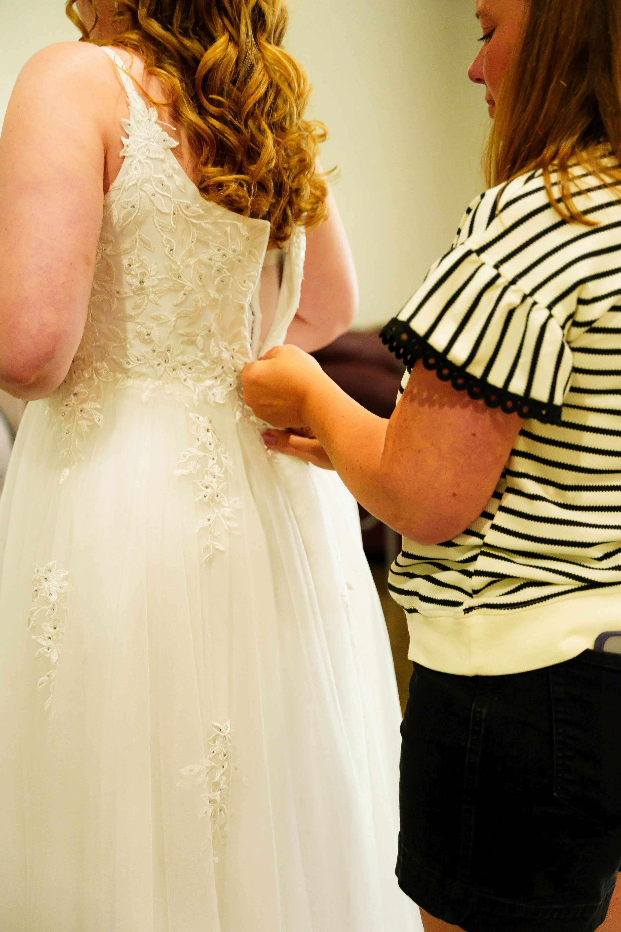 A woman in a striped shirt helps a bride adjust her wedding gown, which features floral embroidery.