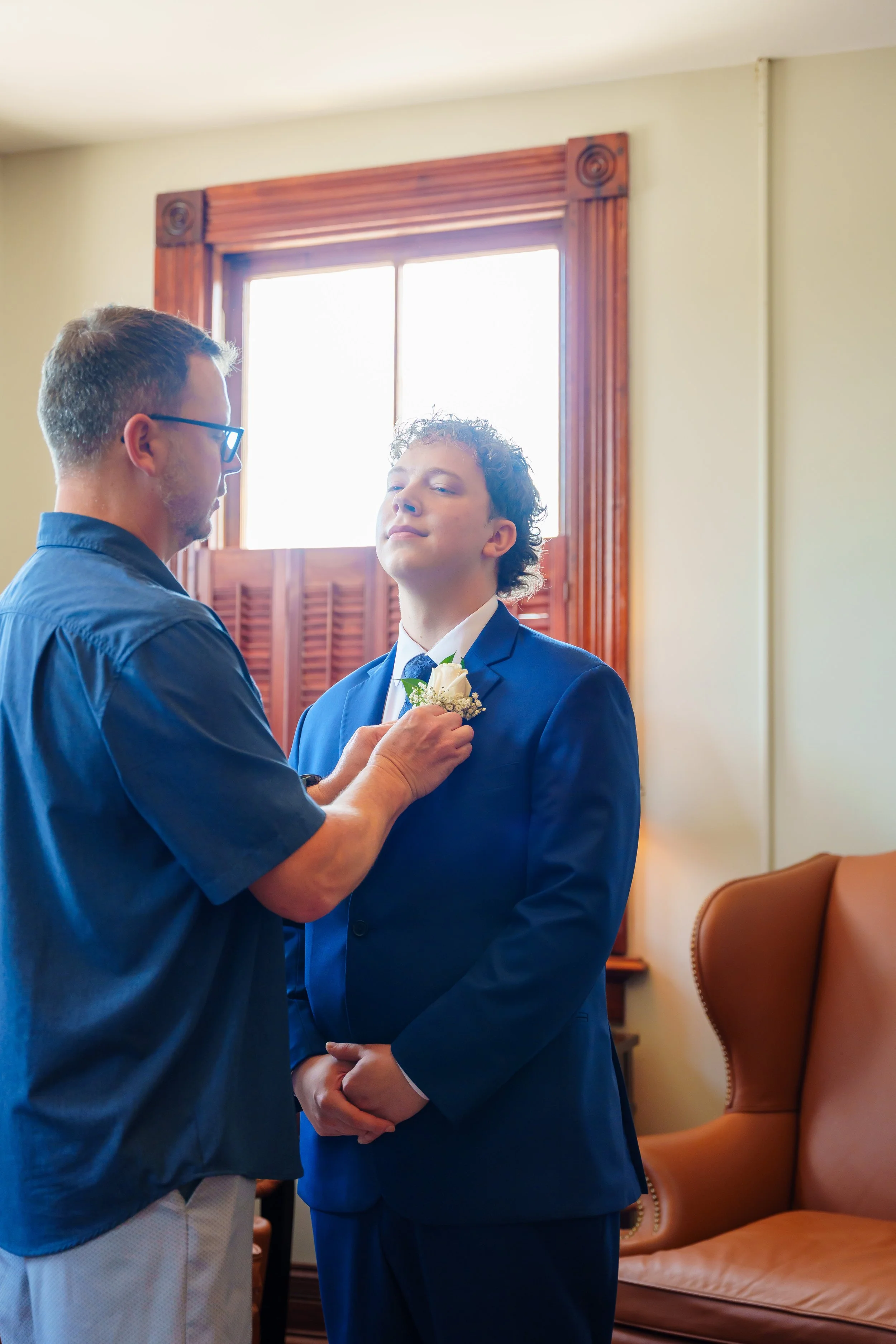 A young man in a dark blue suit with a white shirt stands with closed eyes, receiving a boutonniere from an older man wearing glasses and a navy shirt during a ceremony indoors. A window with wooden framing and shutters is in the background, along wi
