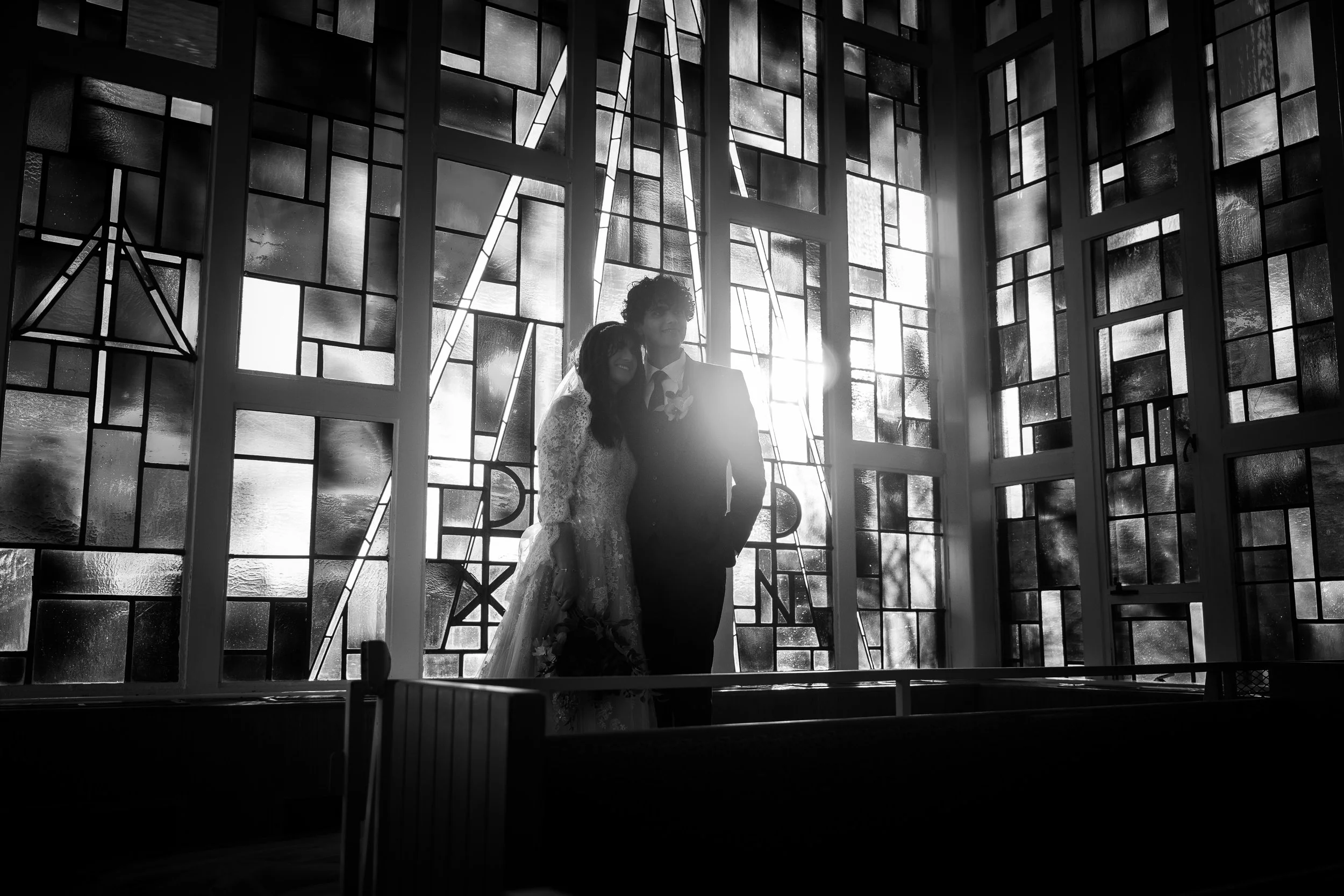 A bride and groom standing together inside a building with large stained glass windows, backlit by sunlight.