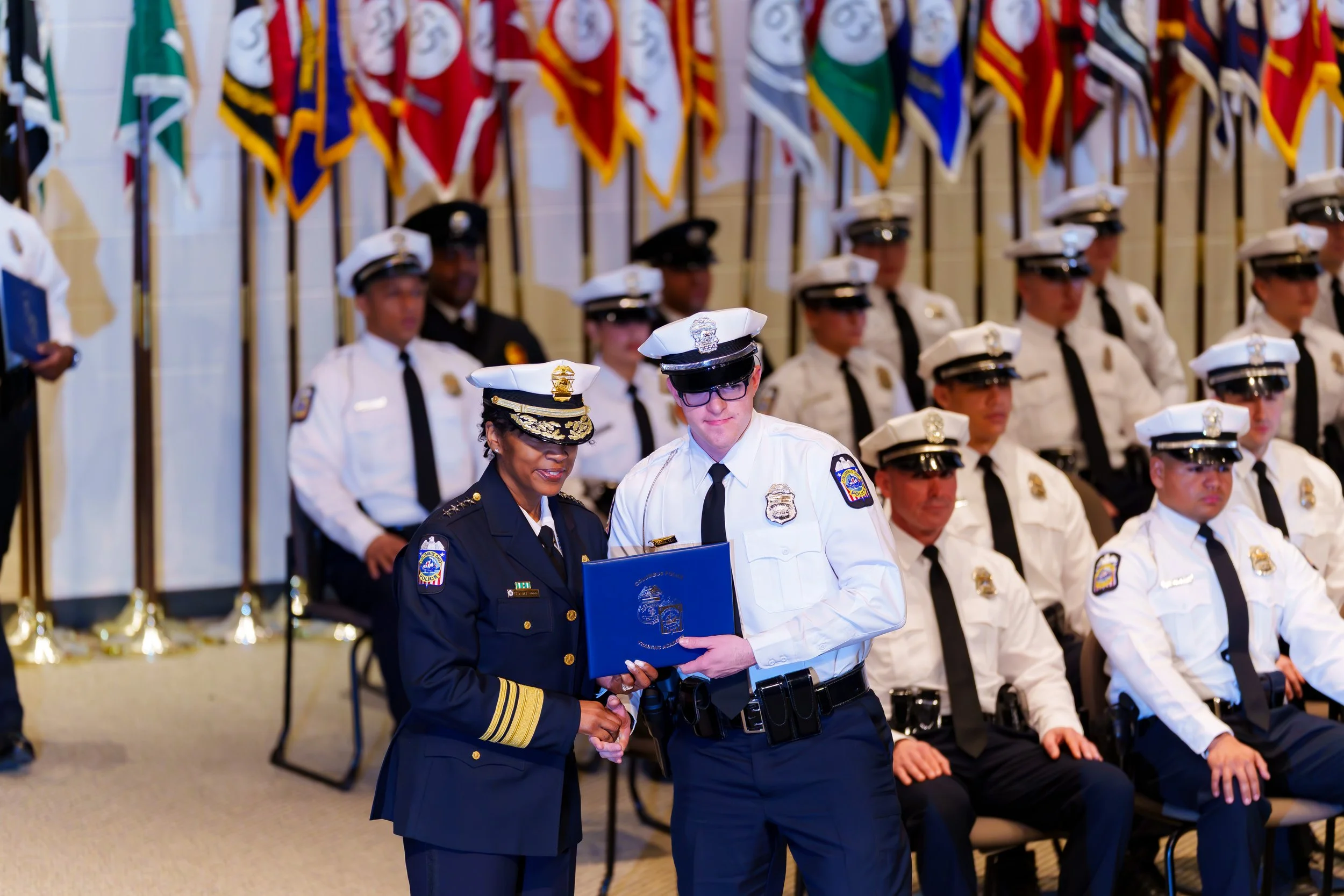 A group of uniformed police officers at an award ceremony, with two officers in the foreground exchanging a certificate, surrounded by flags in the background.