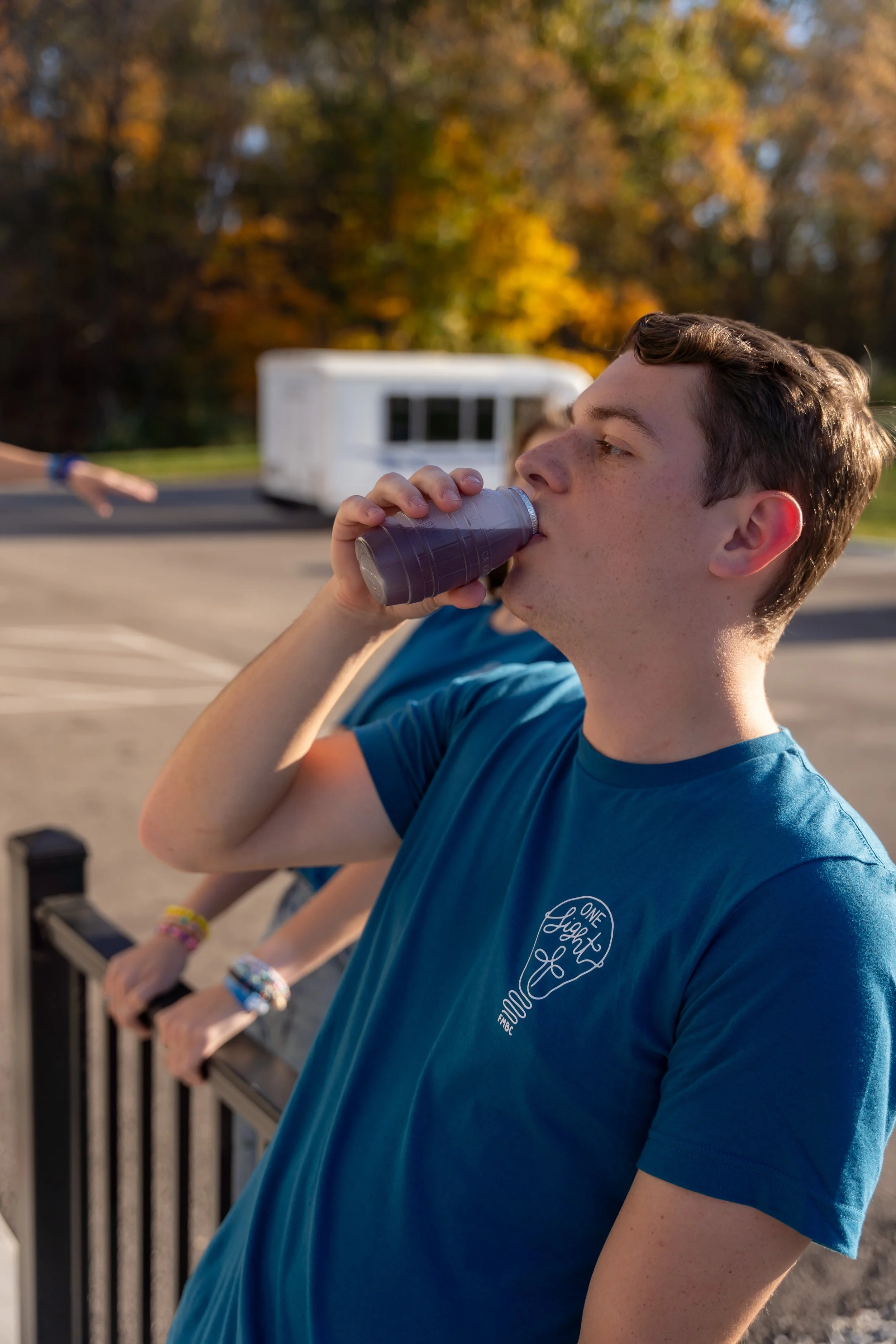 A close-up of a young man with freckles drinking from a plastic bottle with a purple liquid, standing near a railing outdoors with autumn-colored trees in the background.