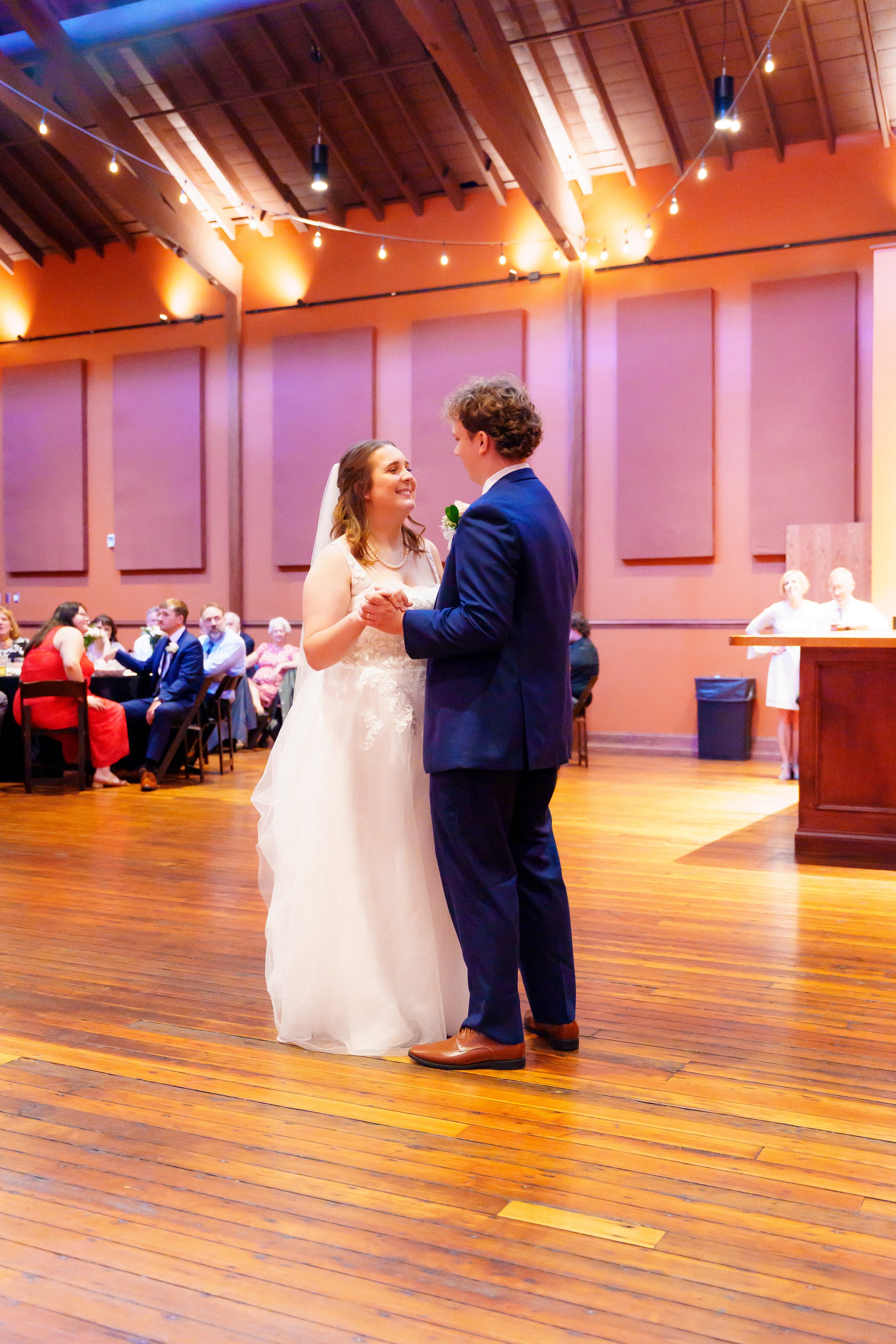 A bride and groom share their first dance at a wedding reception in a decorated indoor venue with string lights, wooden floors, and seated guests watching