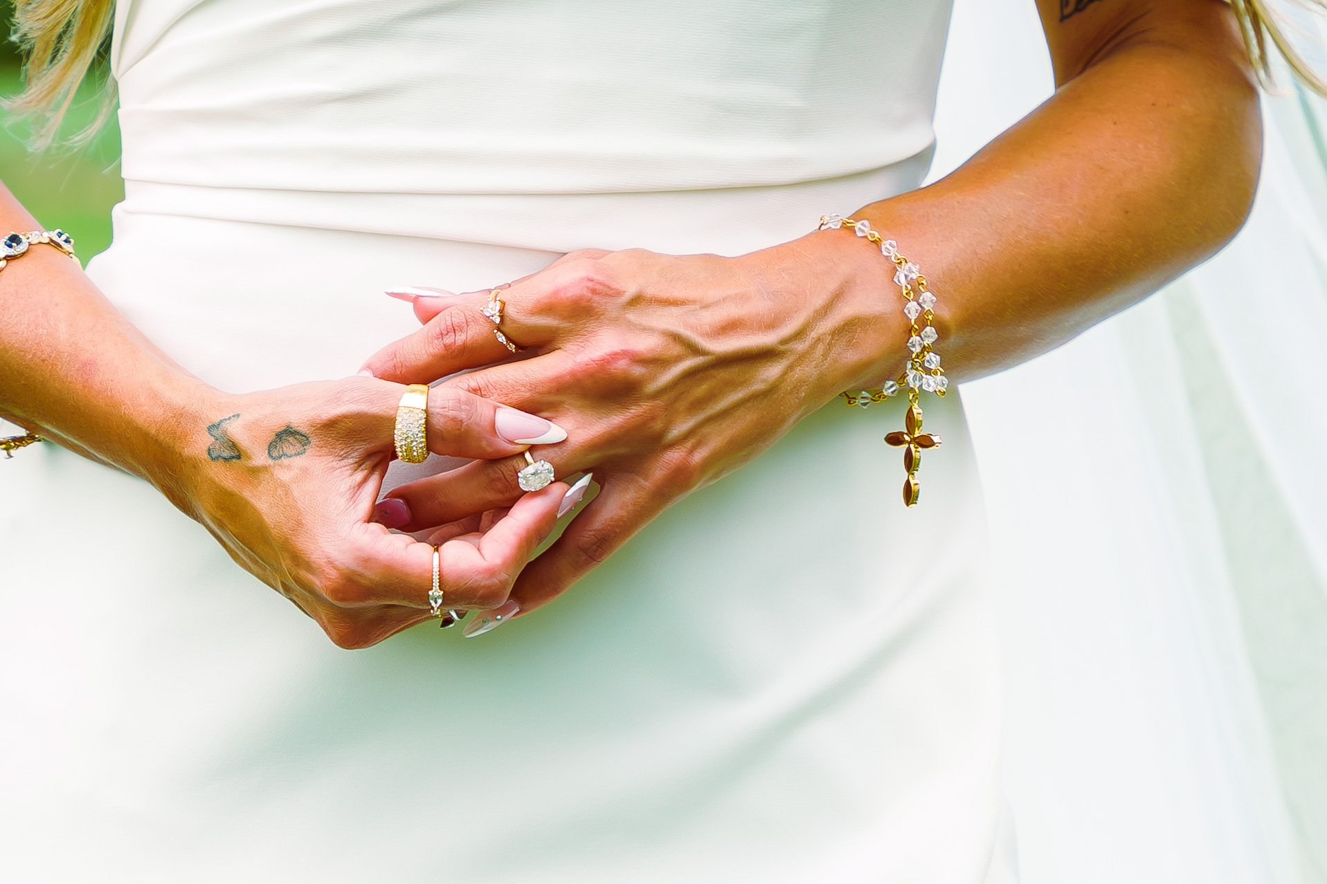 Close-up of a woman's hands, overlapping, with jewelry including rings, bracelets, and tattoos, against a white dress and background.