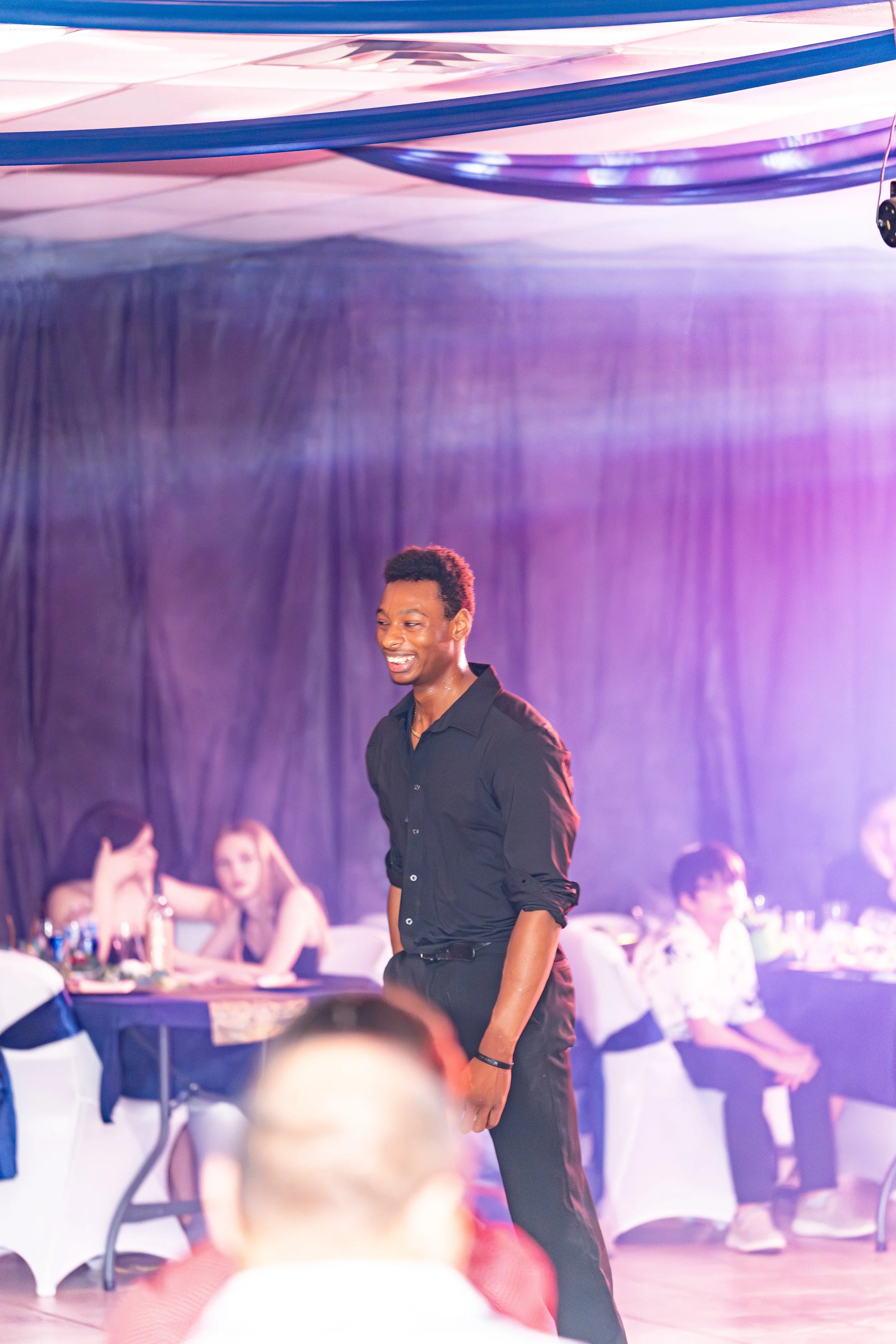 Young man smiling in black shirt, standing at a party or social event with purple lighting, with people sitting at tables in the background.