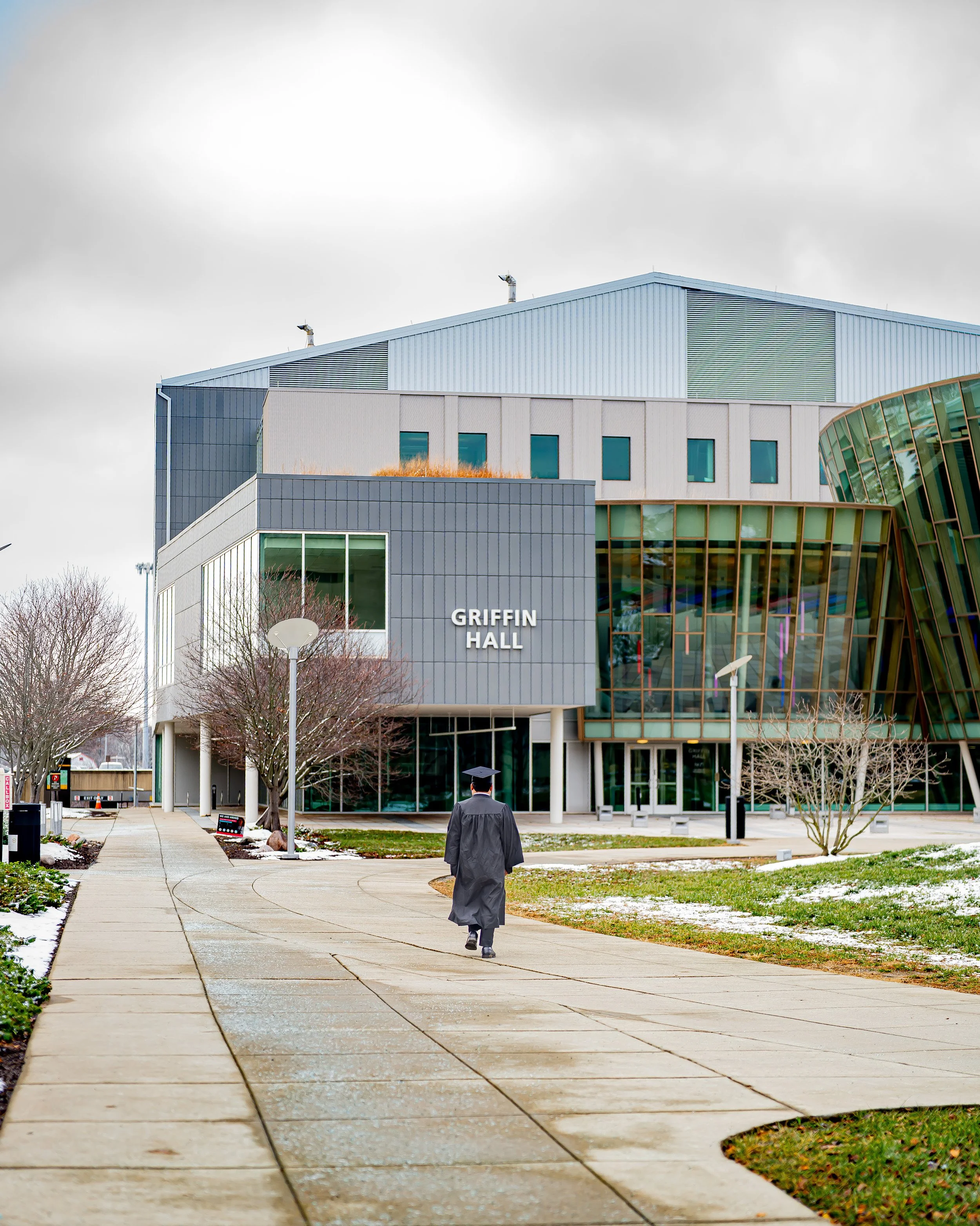 A person in a graduation gown and cap walking on a sidewalk towards Griffin Hall, a modern building with large windows and a unique curved glass section, with leafless trees and cloudy sky in the background.