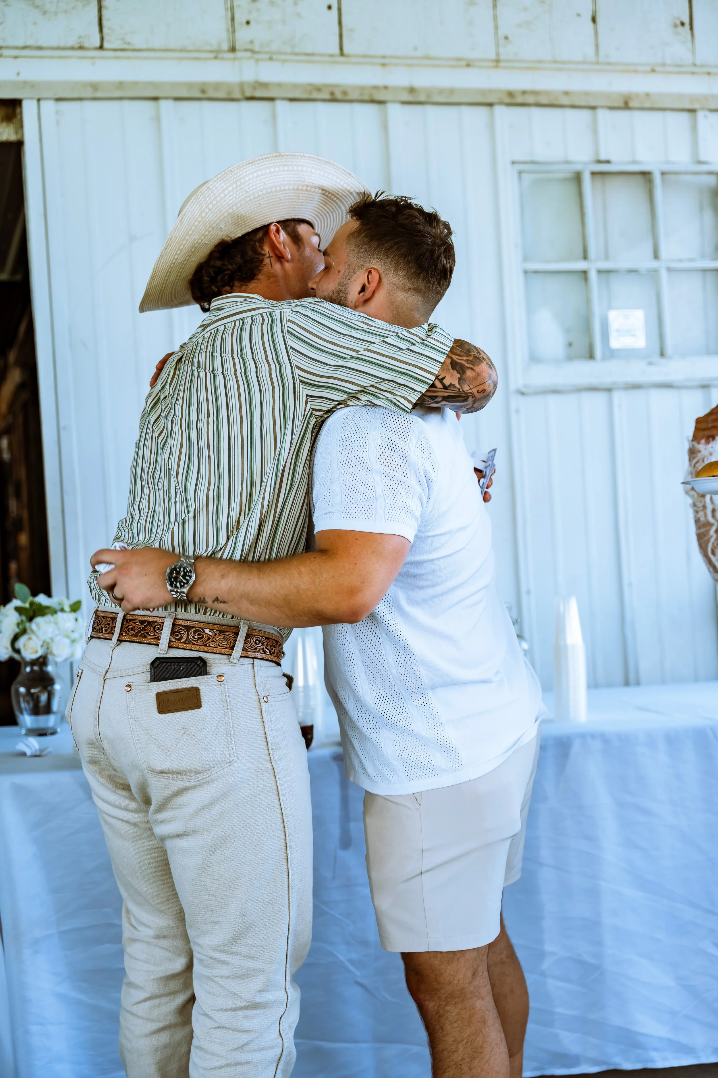 Two men are embracing and kissing in front of a white wooden wall, possibly at a celebration or gathering. One man is wearing a large straw hat and a striped shirt with beige pants, while the other wears a white shirt and white shorts, holding a phon