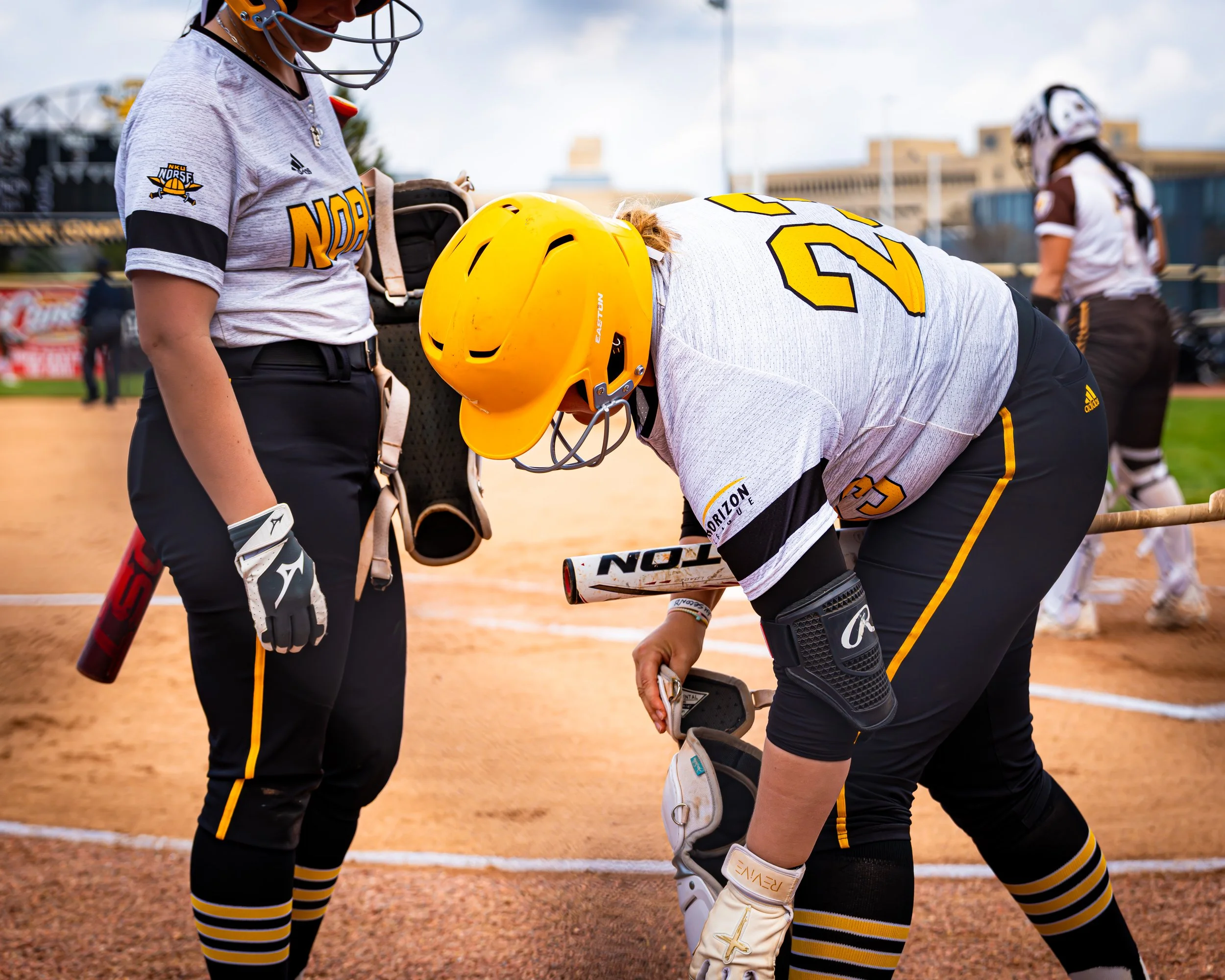 Softball players on the field, with one bending down and another standing with a helmet, in a stadium with buildings in the background.