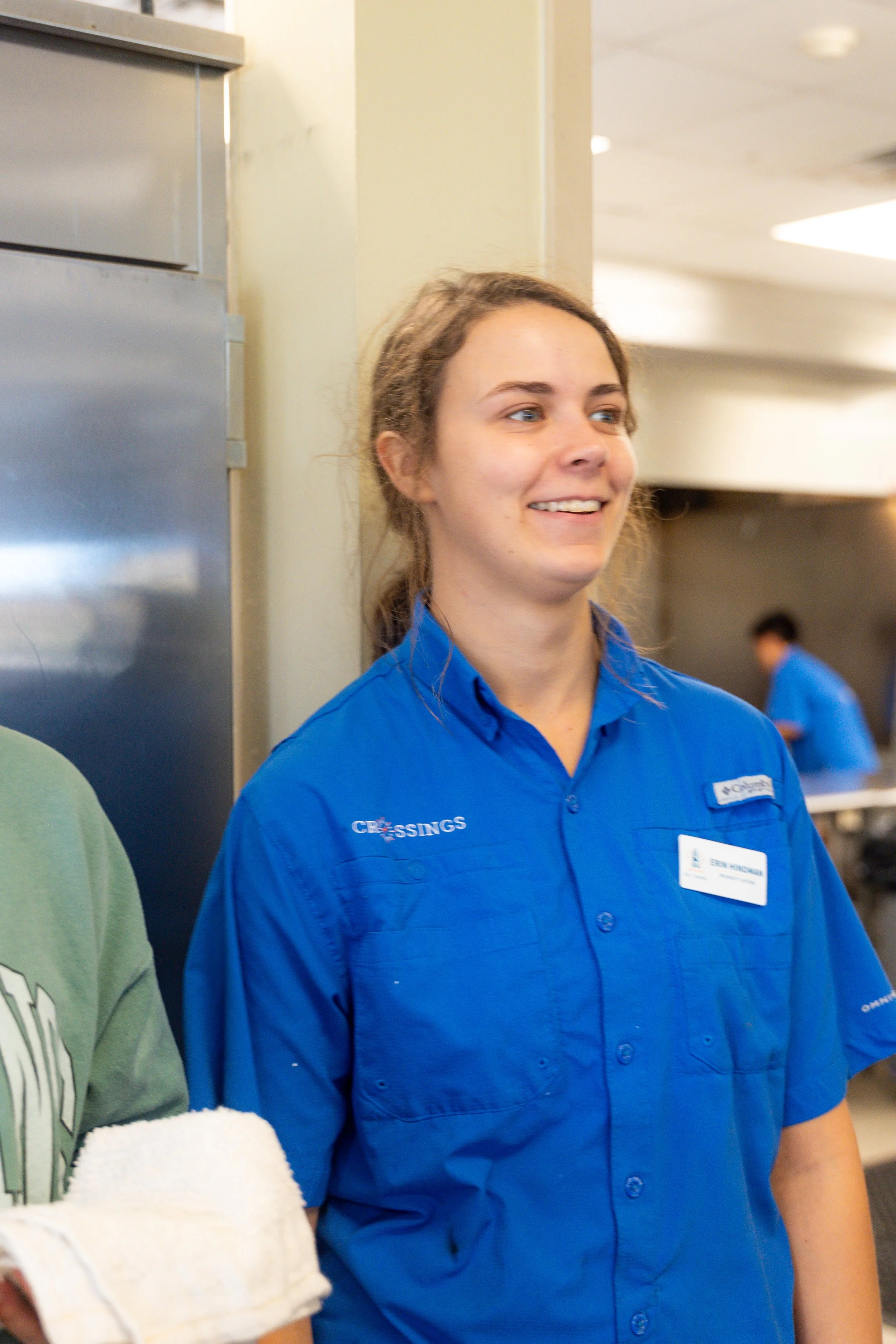 A smiling woman with light brown hair pulled back, wearing a blue shirt with a name tag, standing in what appears to be a commercial or retail setting. Part of another person in a green shirt and an arm with a white towel are visible in the foregroun