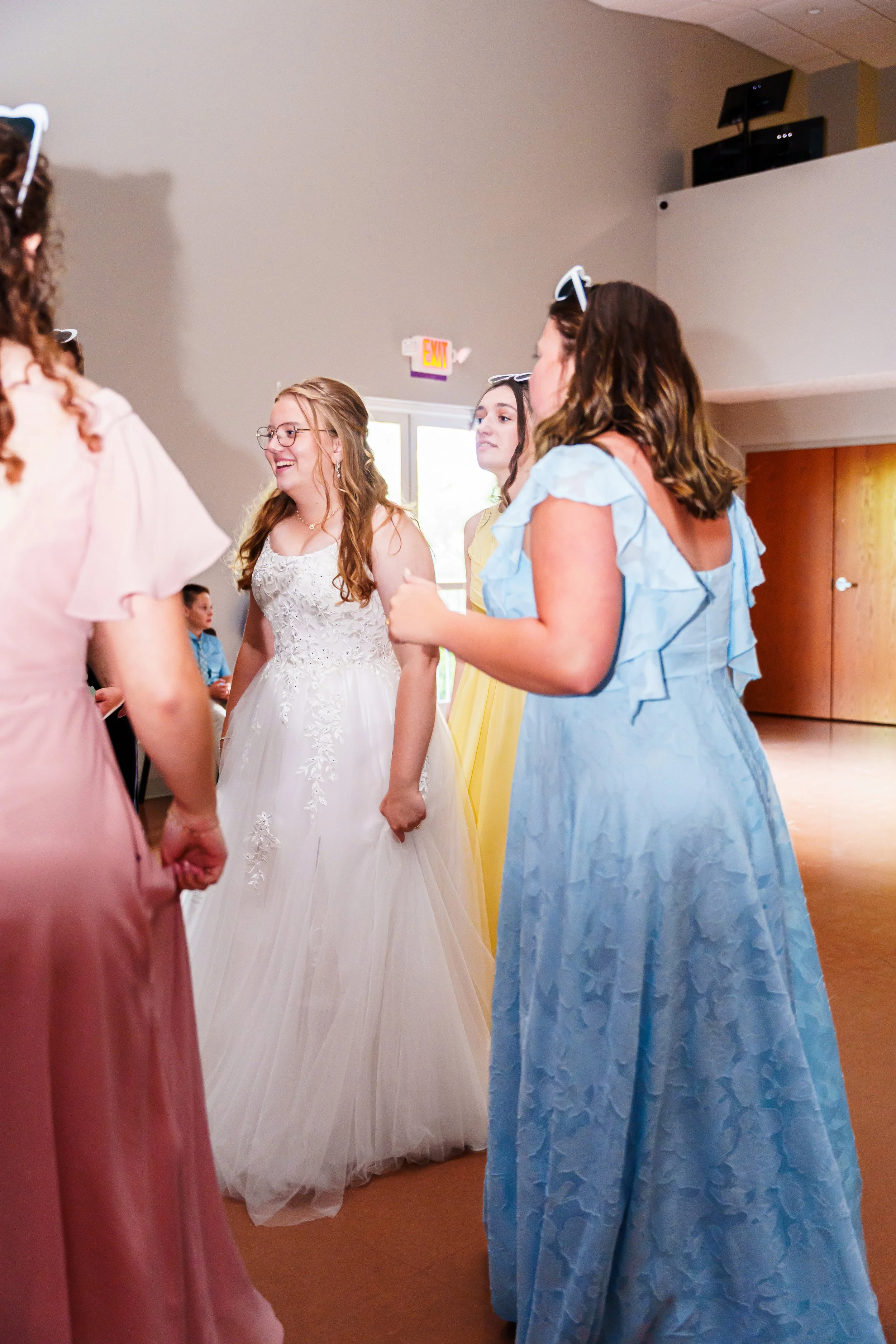 Group of women holding hands, wearing pastel-colored dresses, gathered in a circle, likely at a wedding or celebration, indoors with neutral walls and ceiling, fluorescent lighting, and a door in the background.