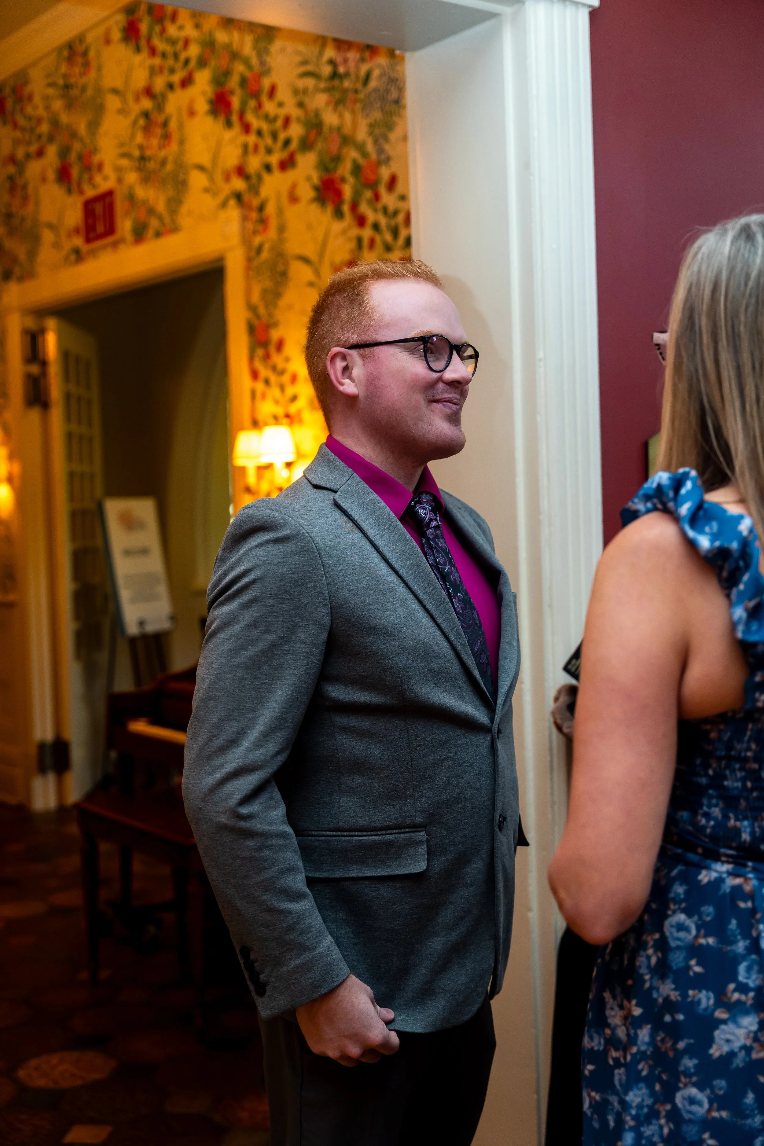 A man wearing glasses, a gray blazer, and a pink shirt is engaged in a conversation with a woman in a blue floral dress in an indoor setting with warm lighting.