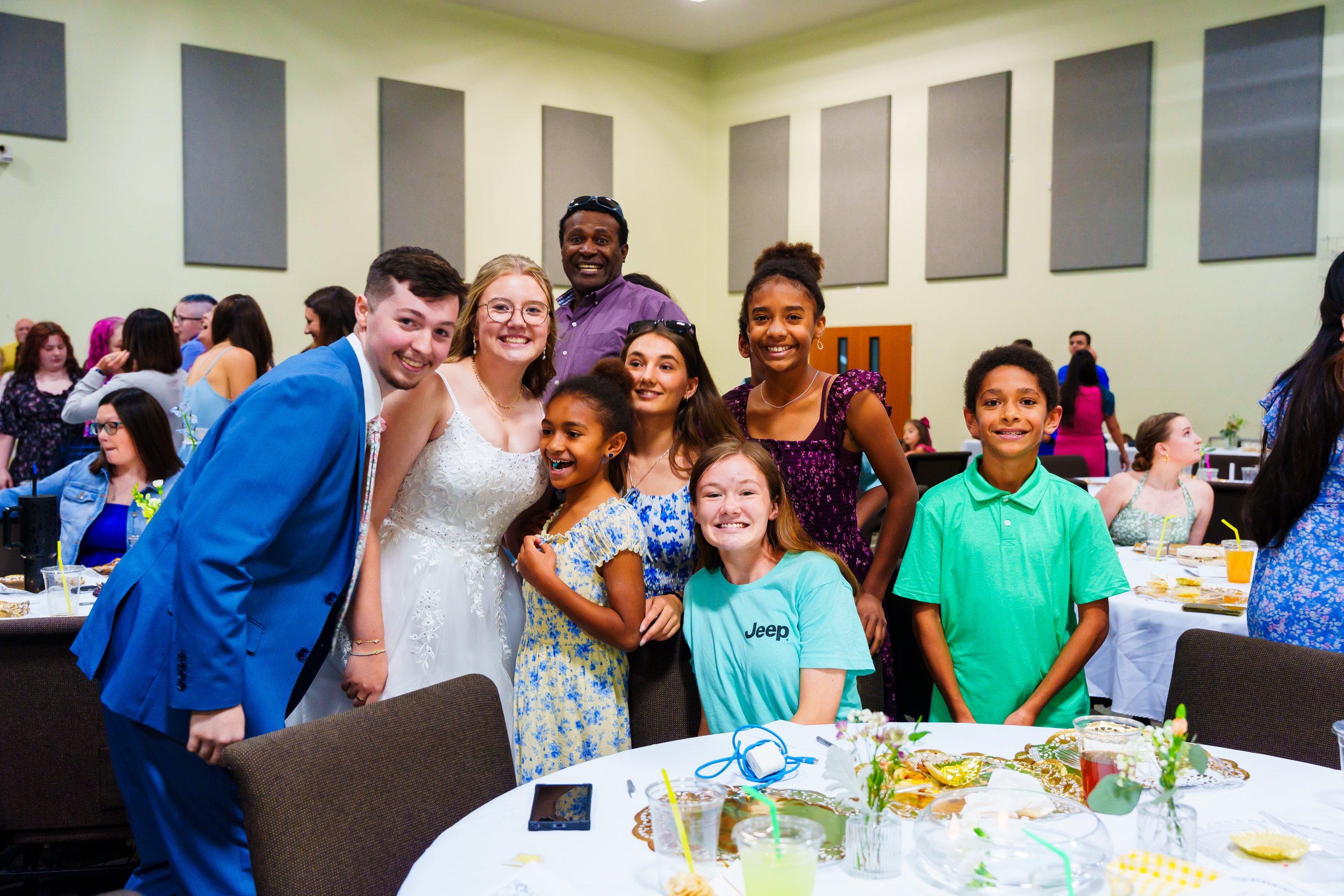 Group of smiling people at a celebration or party, gathered around a table with food and drinks in a decorated room.