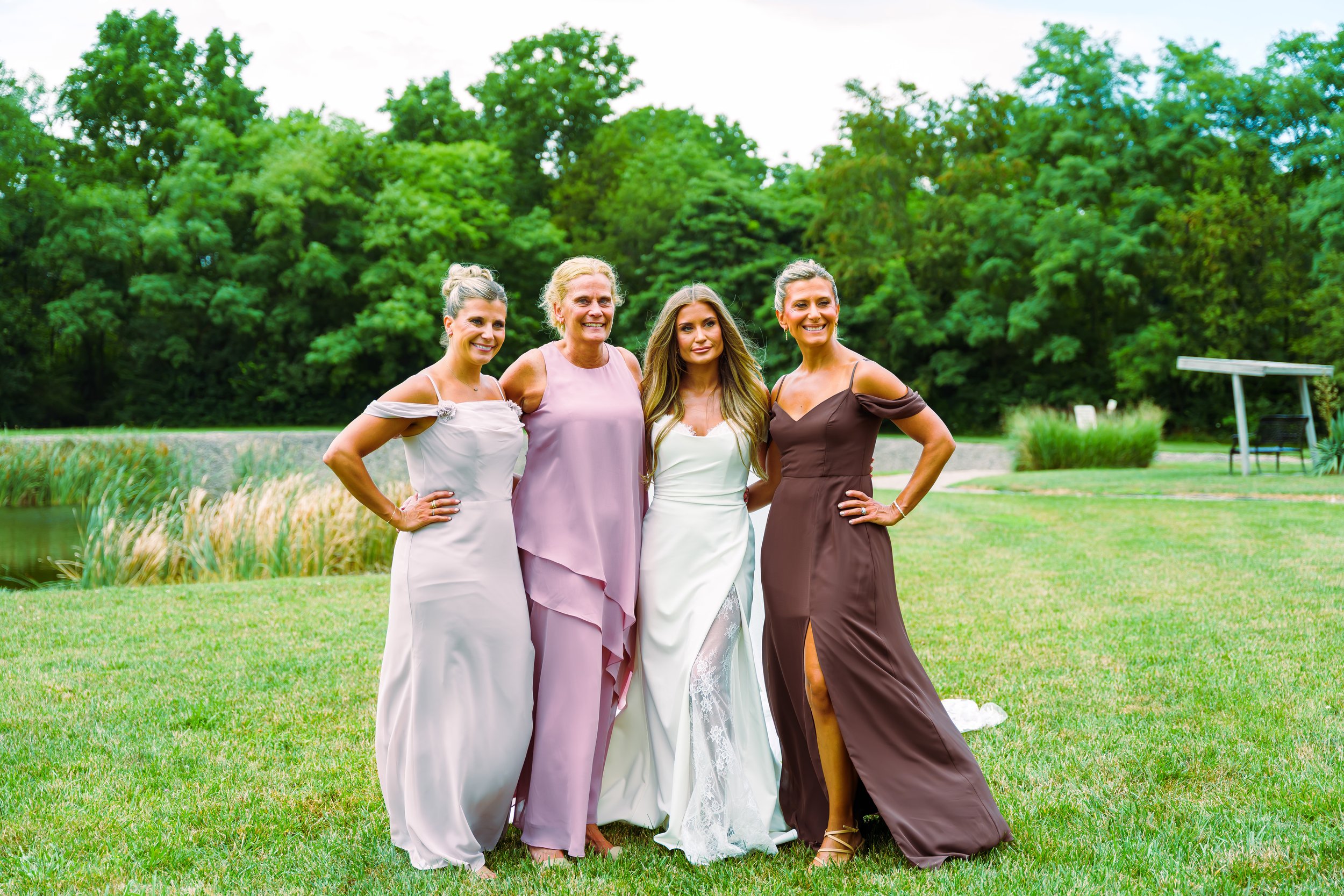Four women in elegant dresses posing together outdoors in front of lush green trees, near a pond or small lake with grass and decorative plants nearby.