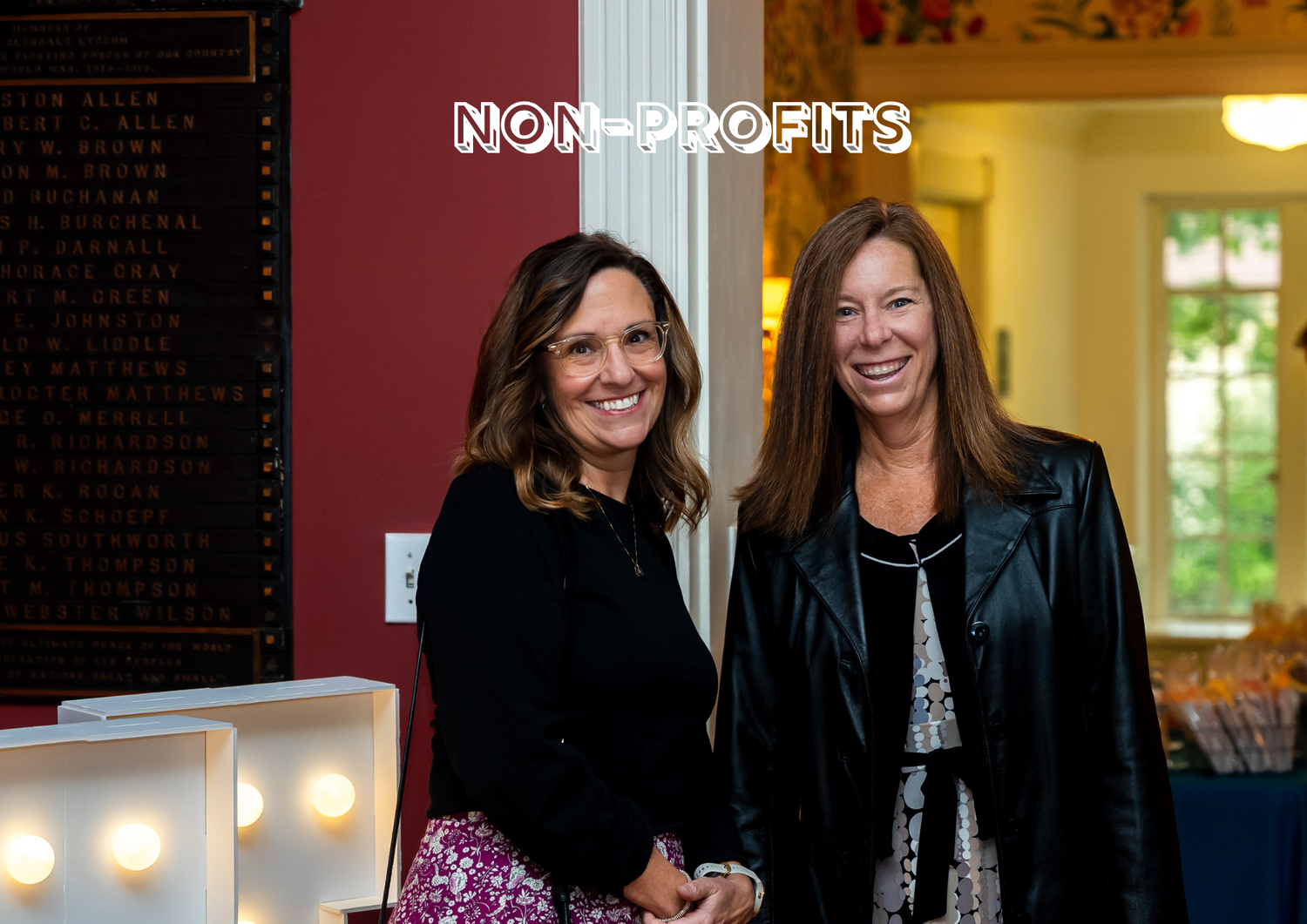 Two women smiling and posing in an indoor setting with a non-profit sign in the background.