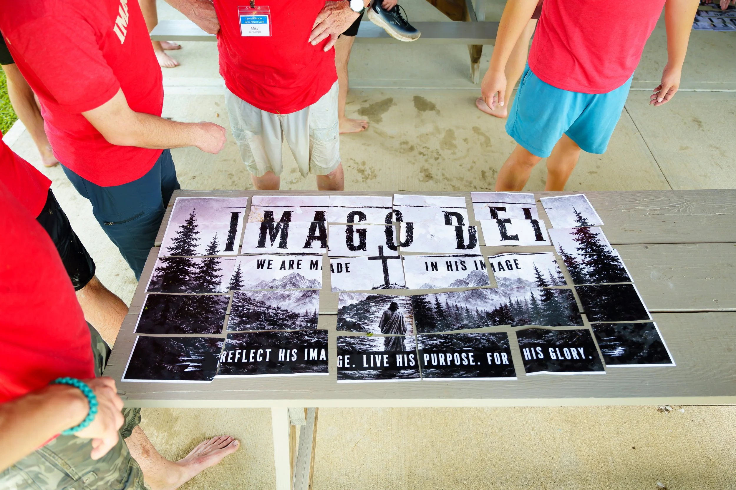 Group of people looking at a display of posters with mountain and forest images, some with the word 'IMAGINE' and messages about reflecting his image, living his purpose, and glory.