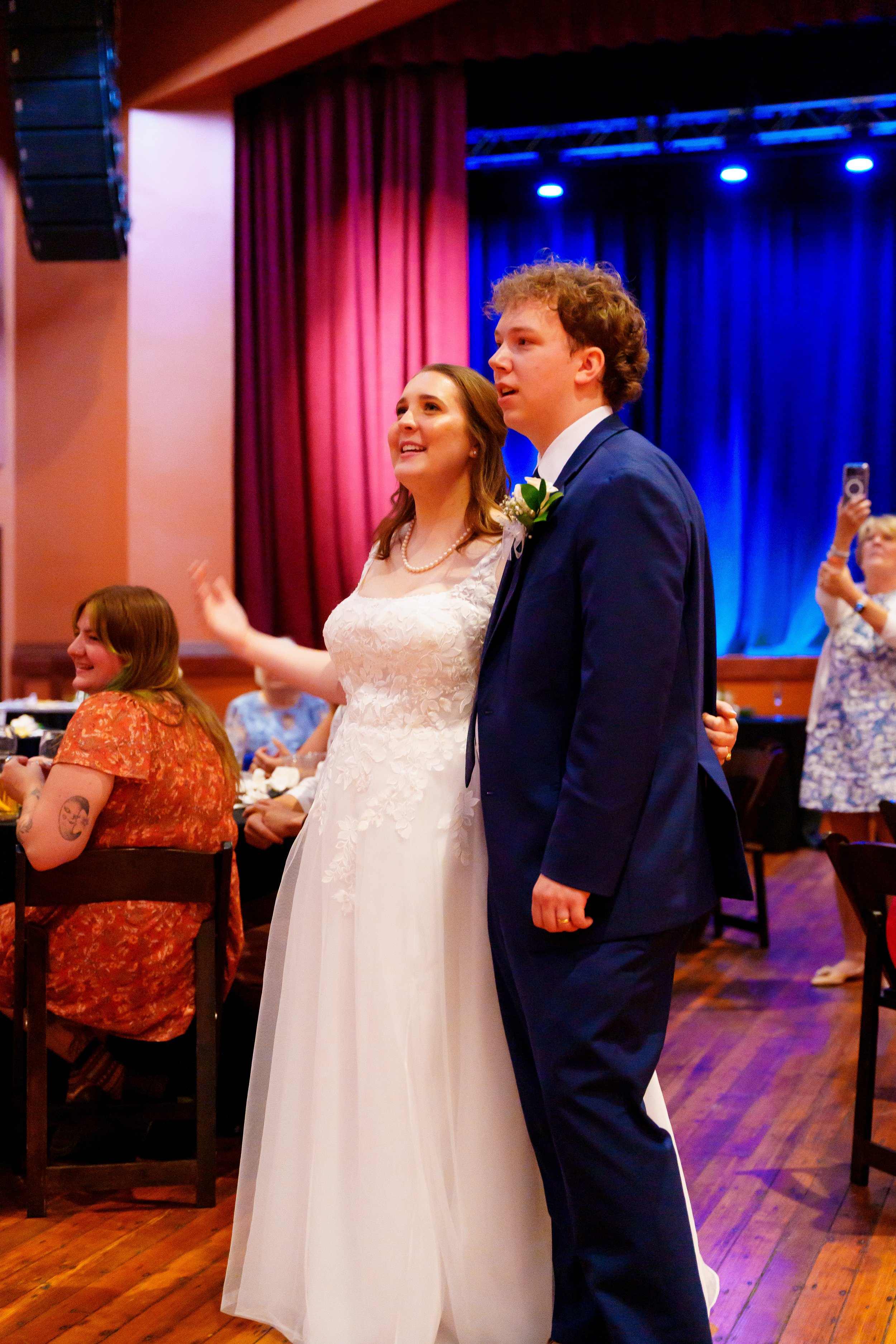 A bride and groom standing together at a wedding reception, with the bride wearing a lace wedding dress and pearl necklace, and the groom in a dark suit with a boutonniere, in a warmly lit venue with red and blue curtains in the background.