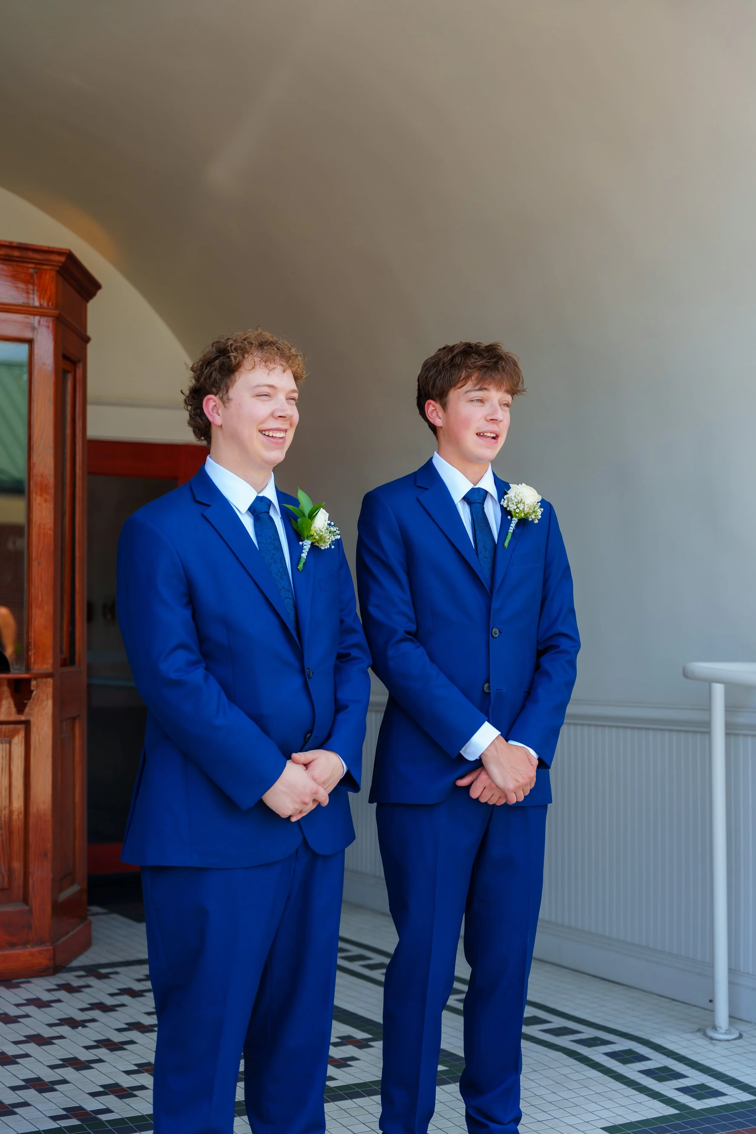 Two young men in matching blue suits, white shirts, and blue ties stand side by side indoors, each with a white floral boutonniere on their lapels. They are smiling and looking slightly to the side.