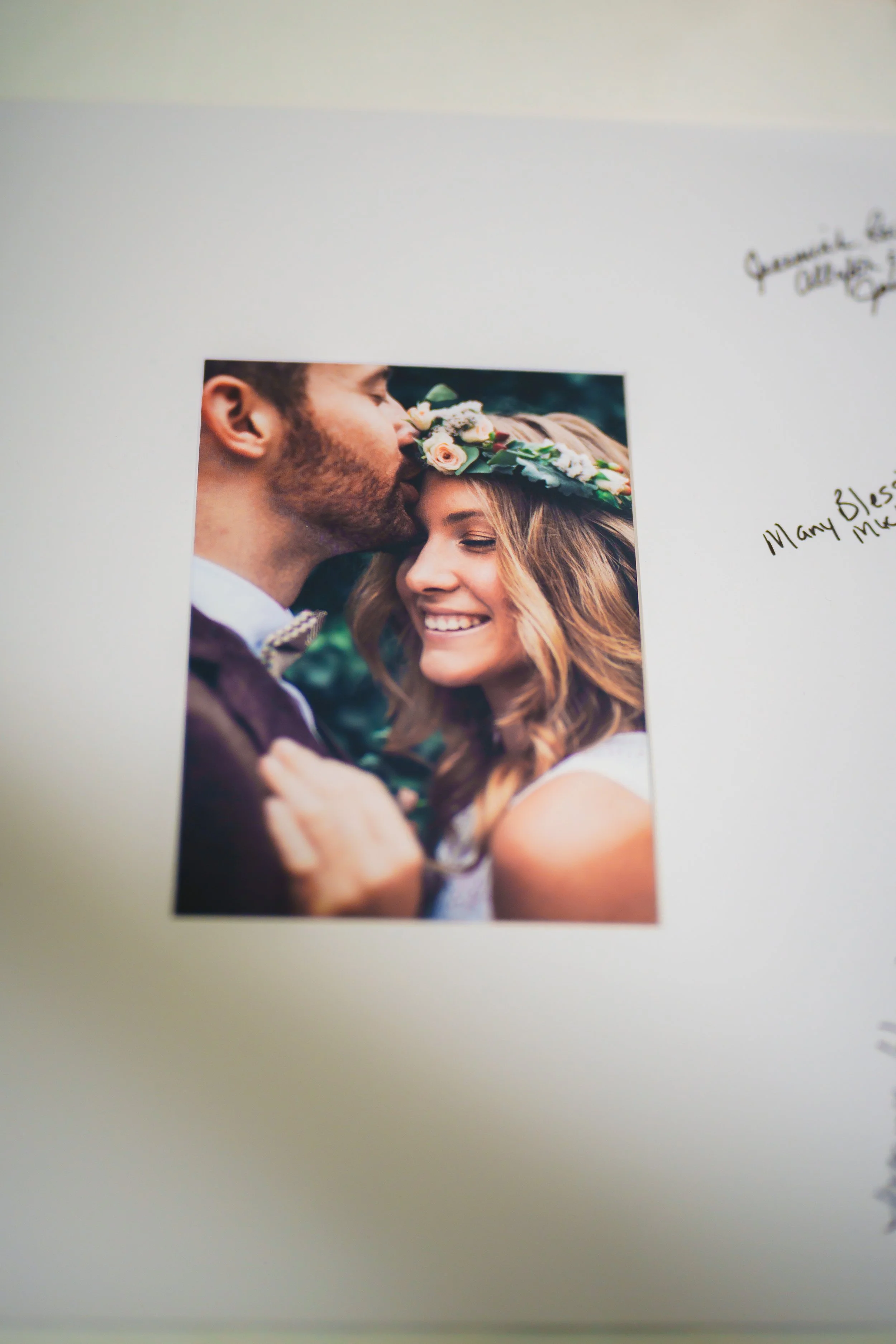 A printed photo of a couple, with the man kissing the woman's forehead, both smiling with closed eyes, outdoors with blurred greenery. The woman has a flower crown. There are handwritten notes on the paper around the photo.