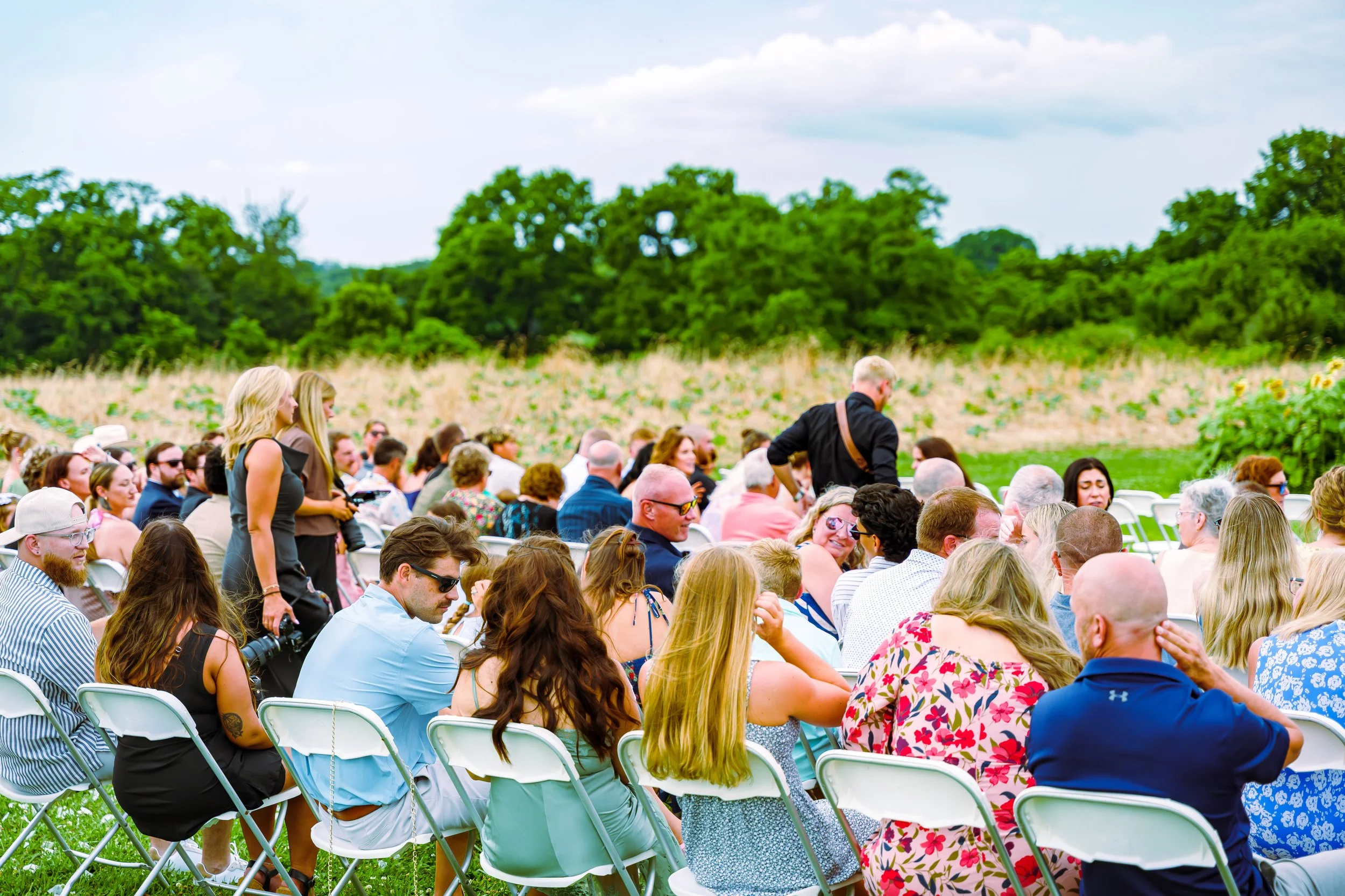 A large outdoor gathering of people seated on white folding chairs in a field with green trees and a partly cloudy sky in the background.