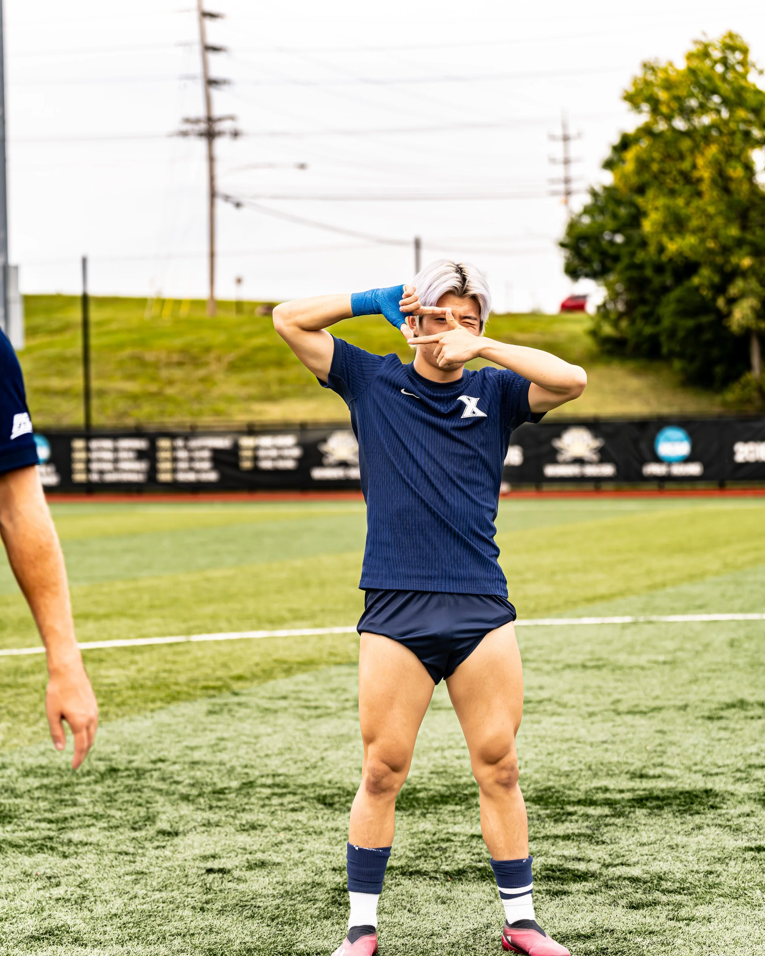 A young male soccer player with short, blonde hair wearing a blue sports shirt, dark shorts, long blue and white socks, and pink cleats, stands on a green soccer field making a playful gesture with his fingers around his eyes. He is wearing a blue wr