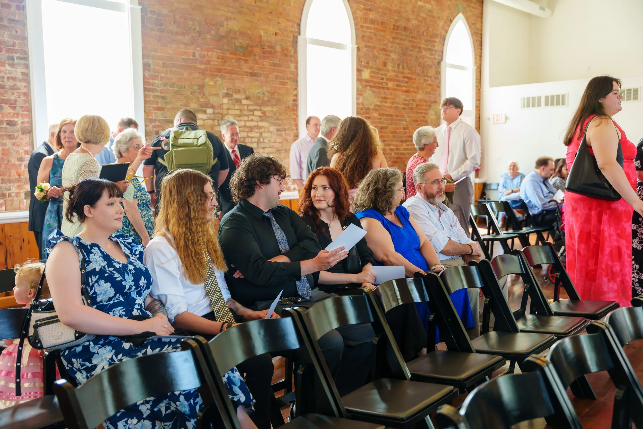 A diverse group of people seated and standing in a spacious room with brick walls and tall arched windows, attending a formal event or ceremony.