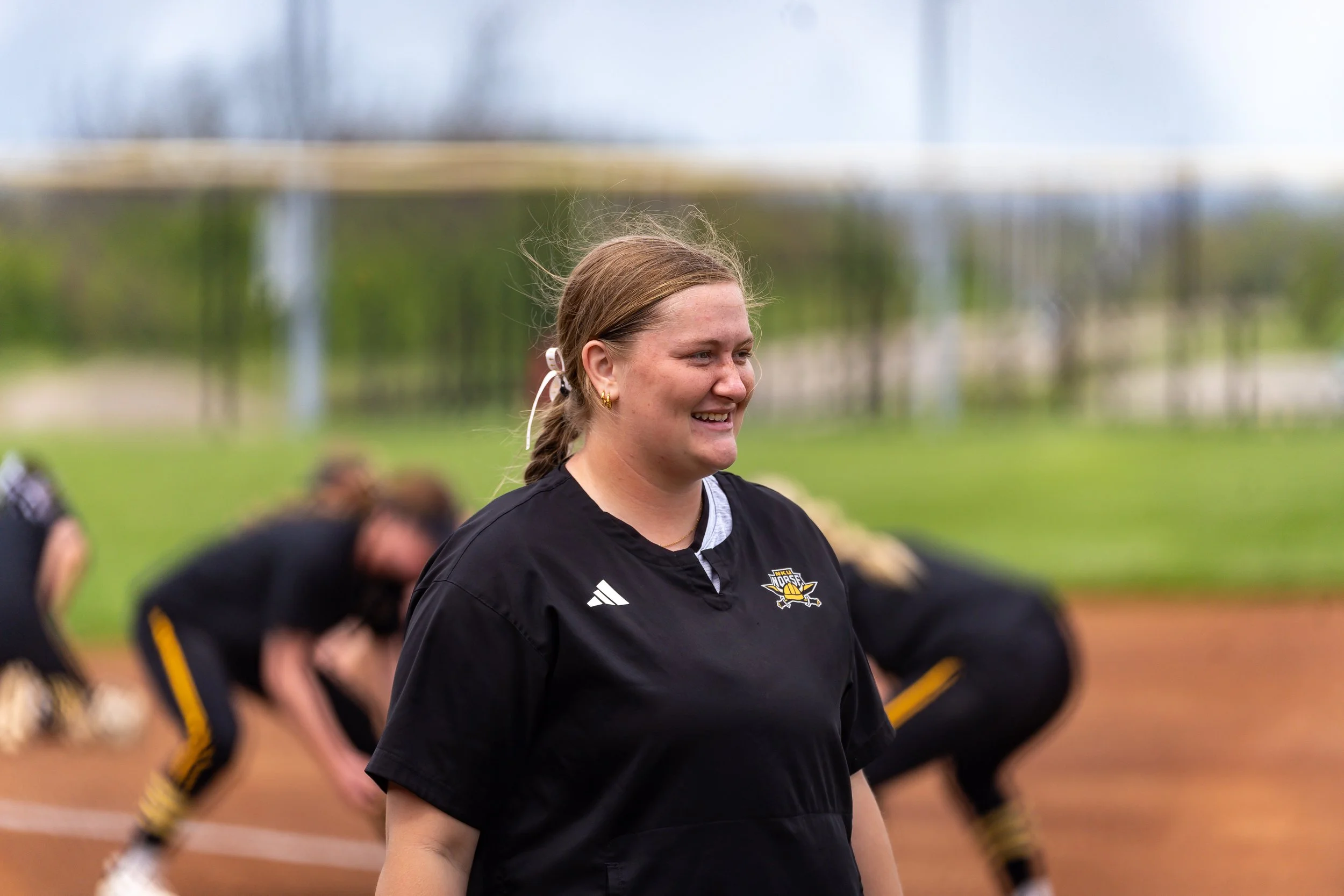 A woman with brown hair tied back, wearing a black sports jersey with a logo, smiling on a sports field with a blurred group of people in similar attire in the background.