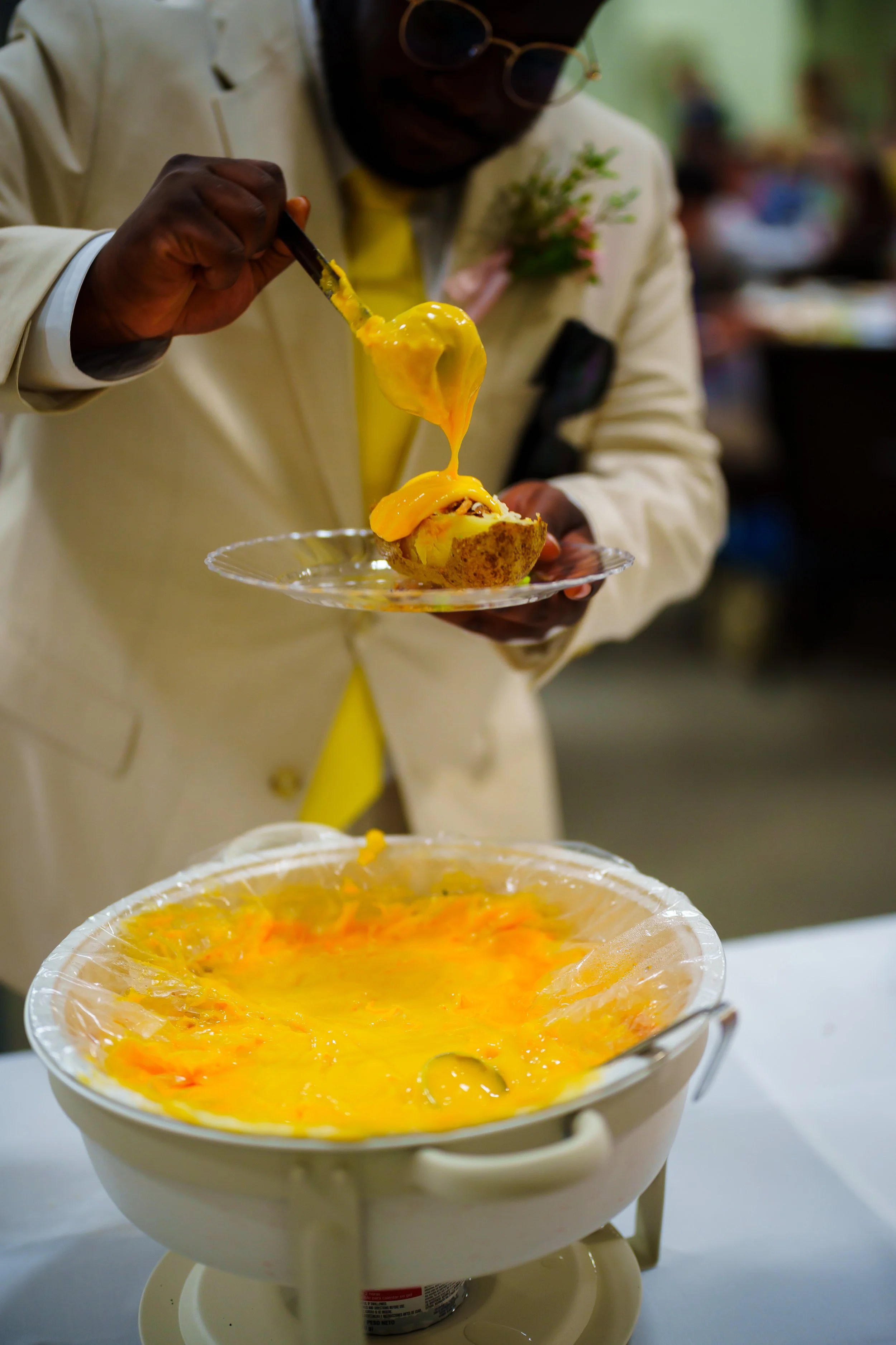 Man with glasses and a white jacket serving melted cheese dip into a bowl below with a spoon. The bowl is lined with plastic wrap and contains an orange-colored dish, possibly a cheese or vegetable dip.