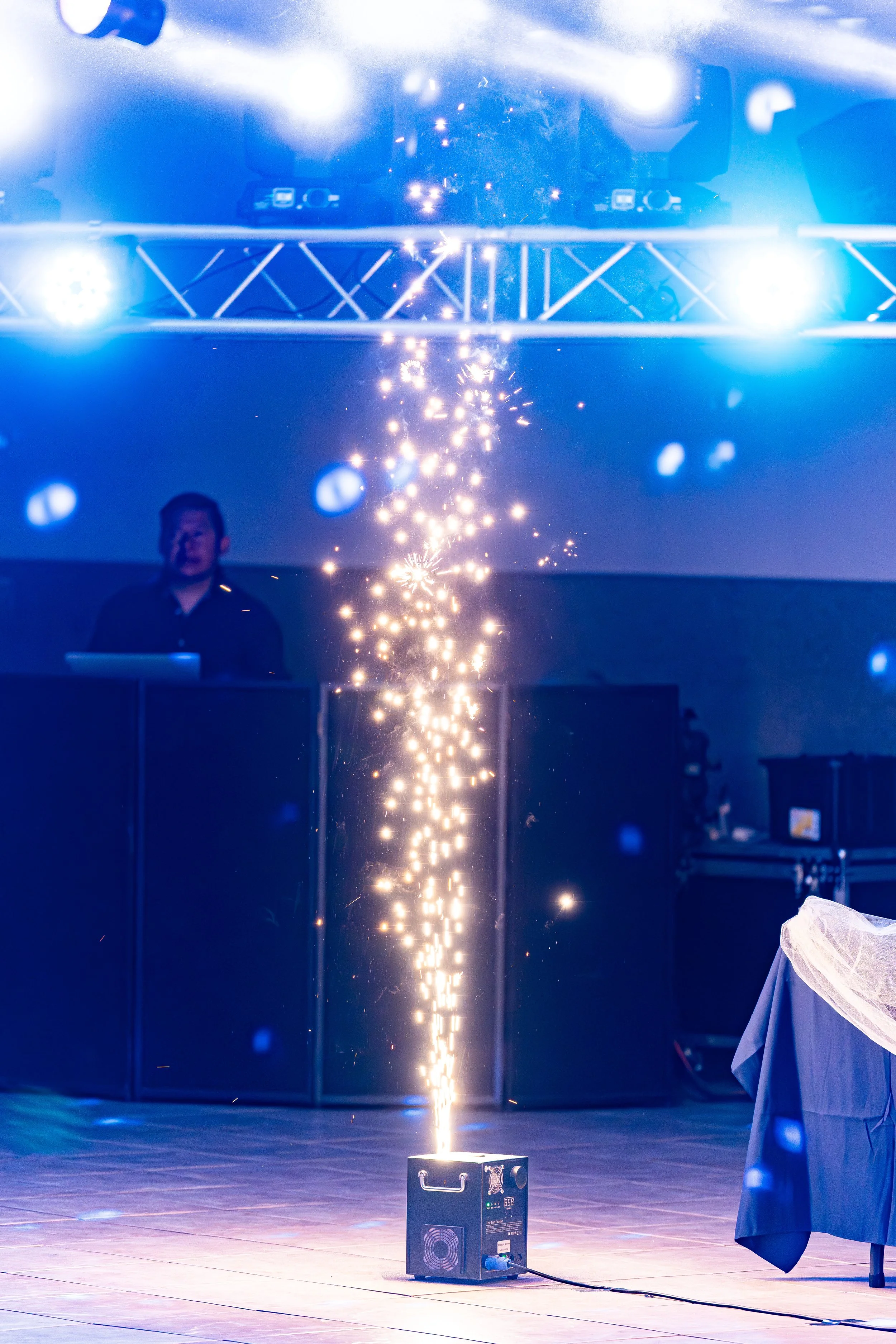 A sparkler or pyrotechnic device producing bright sparks displayed on a stage with blue lighting, a person in dark clothing standing nearby, and equipment on the floor.