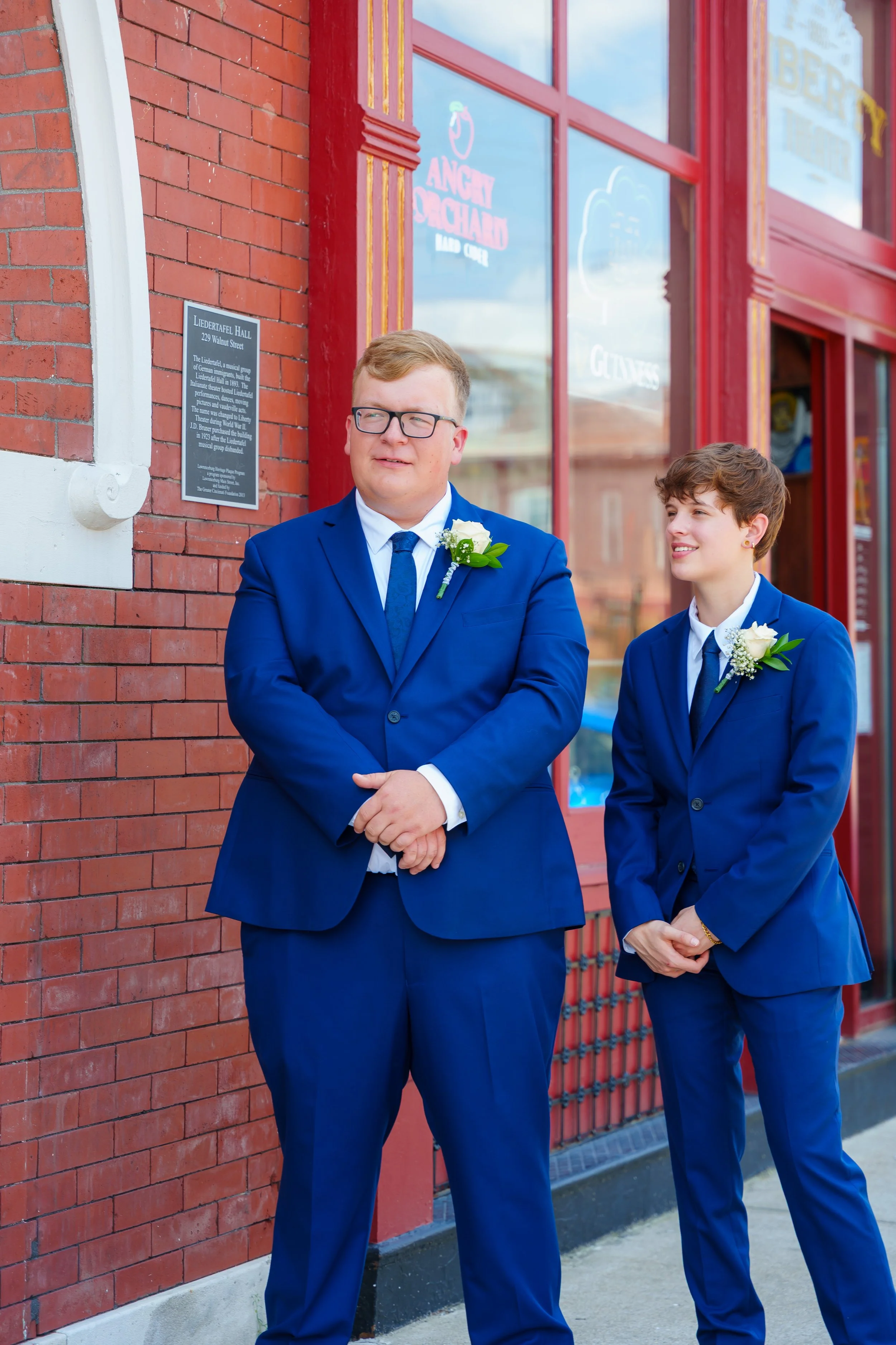 Two people dressed in blue suits with white shirts and floral boutonnieres stand outside a brick building with red-framed windows and a plaque on the wall.