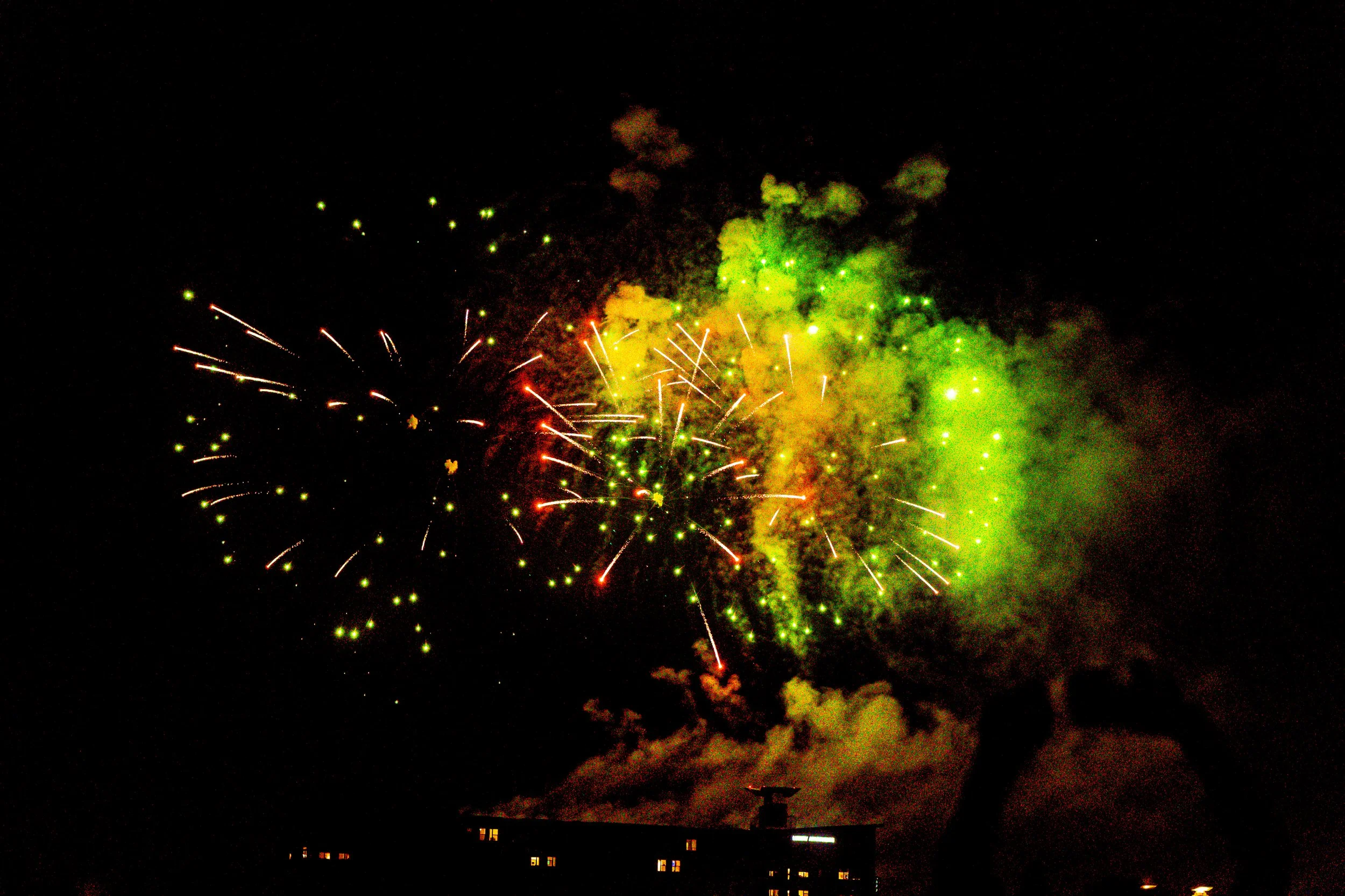 Colorful fireworks exploding in the night sky with green and red hues, displaying sparks and smoke.