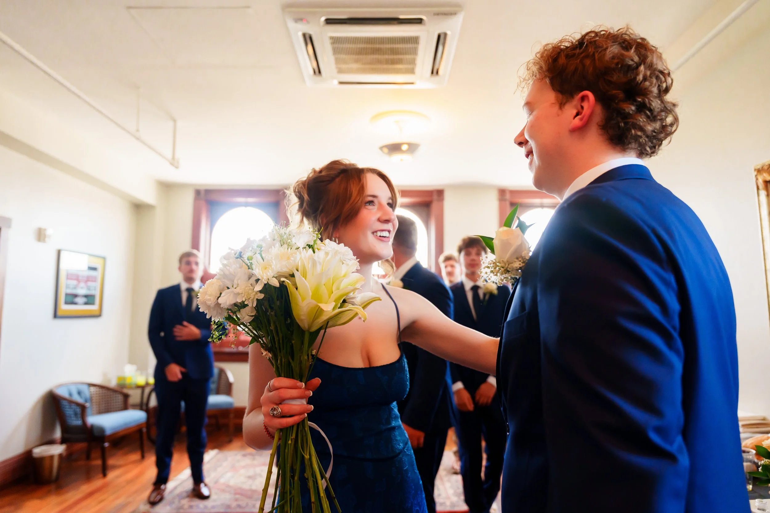 A bride smiling and holding a bouquet of white flowers while looking at a groom during a wedding ceremony in an indoor setting with other people in the background.