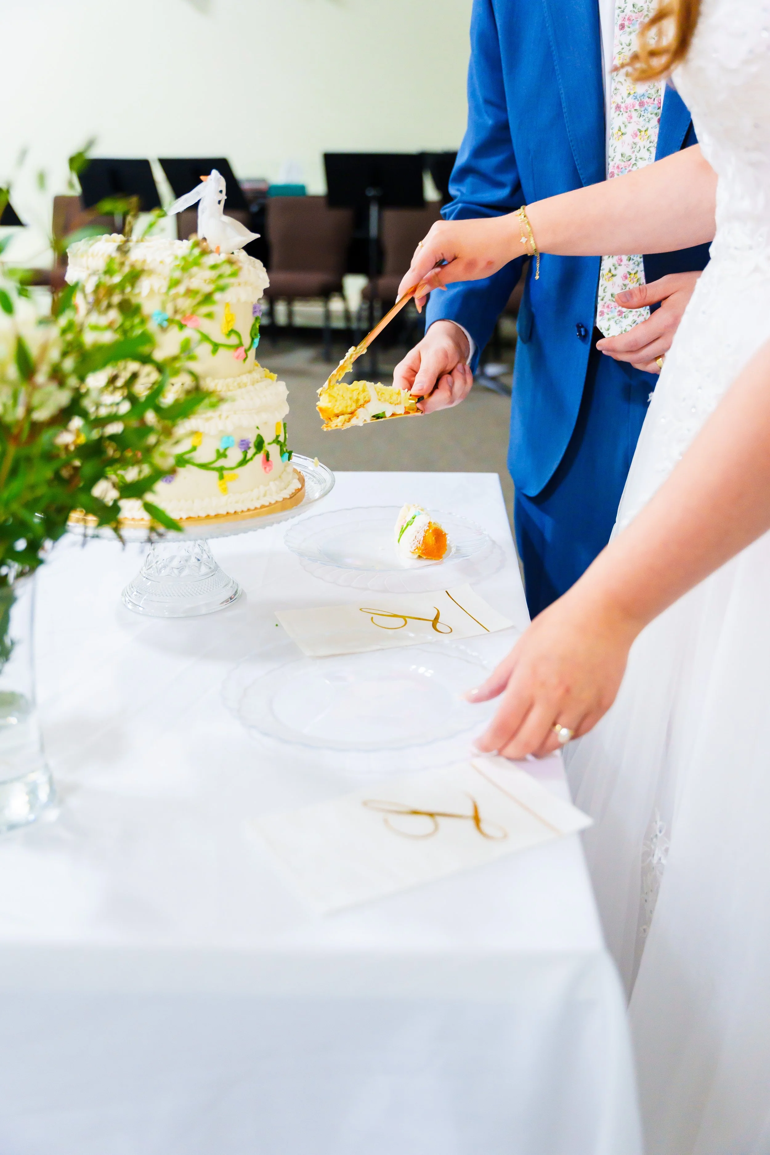People cutting a wedding cake with white frosting and colorful decorations, in a room with empty chairs in the background.