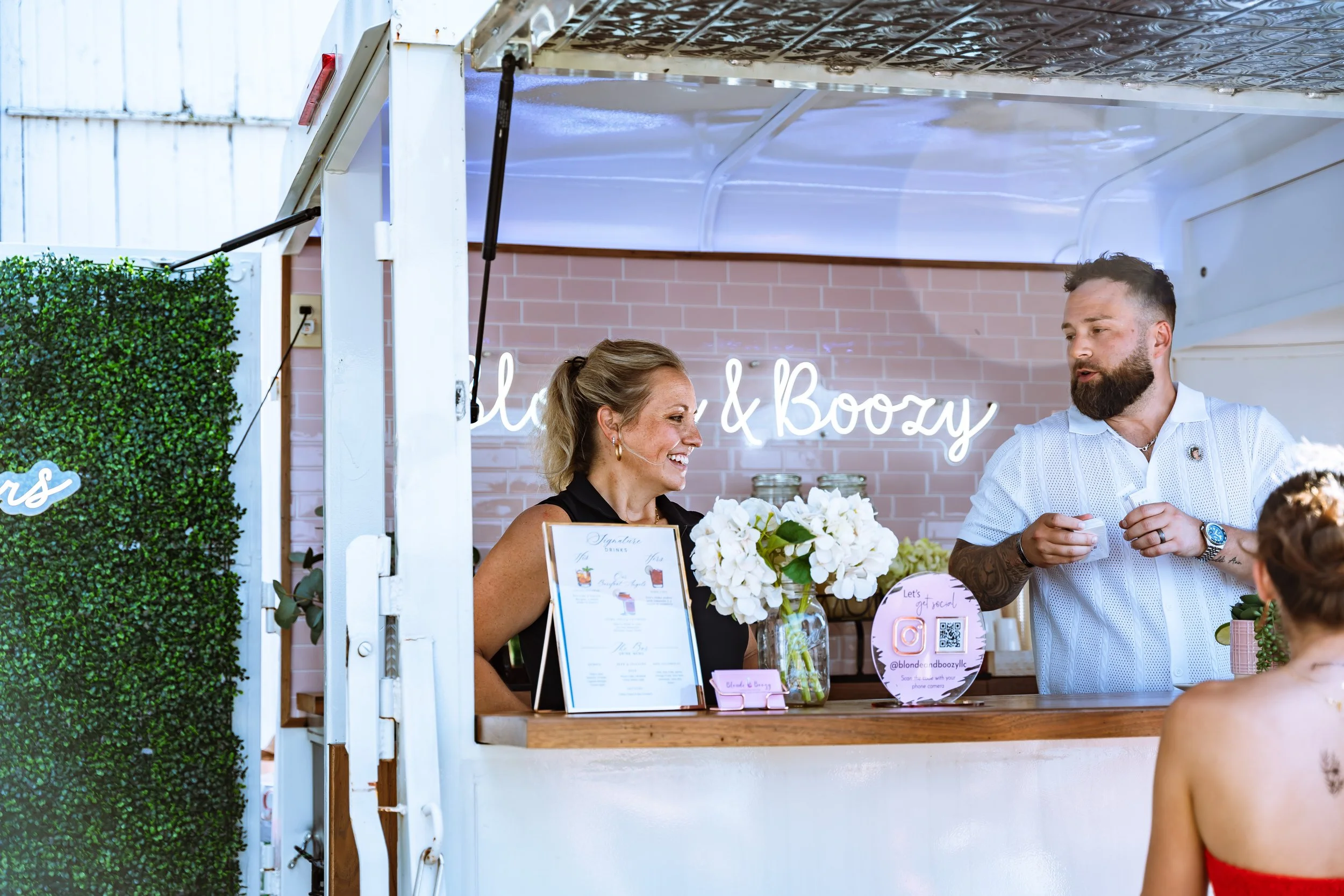 A woman and a man standing behind a food or drink stand at an outdoor event. The woman is smiling, holding a bouquet of white flowers, and the man is talking to someone. The stand has a menu, a sign with an Instagram handle, and various jars and deco