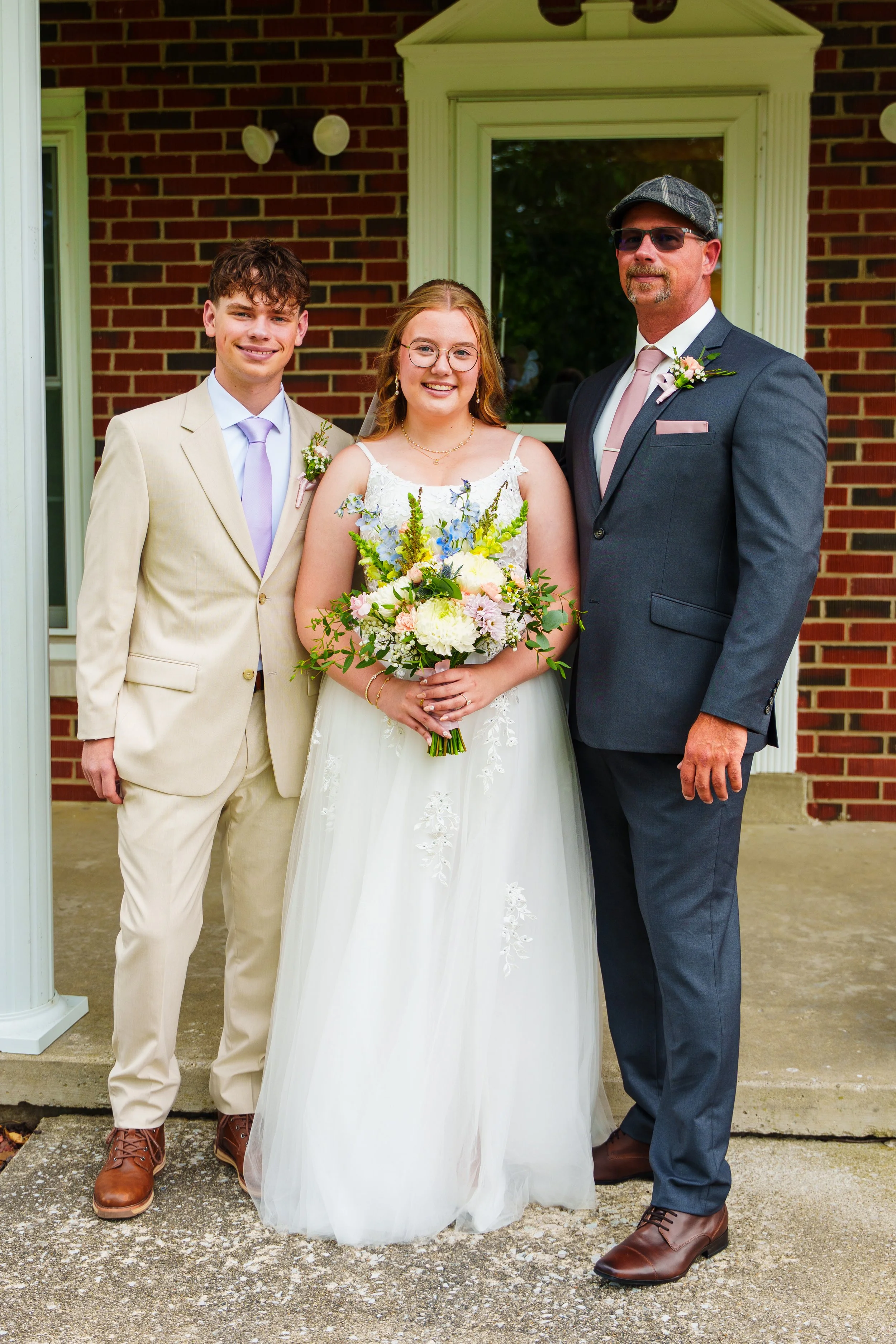 Three people in formal attire standing outside in front of a brick building: a young man in a beige suit with a purple tie, a young woman in a white wedding dress holding a bouquet, and an older man in a dark gray suit with a pink tie and hat, all sm
