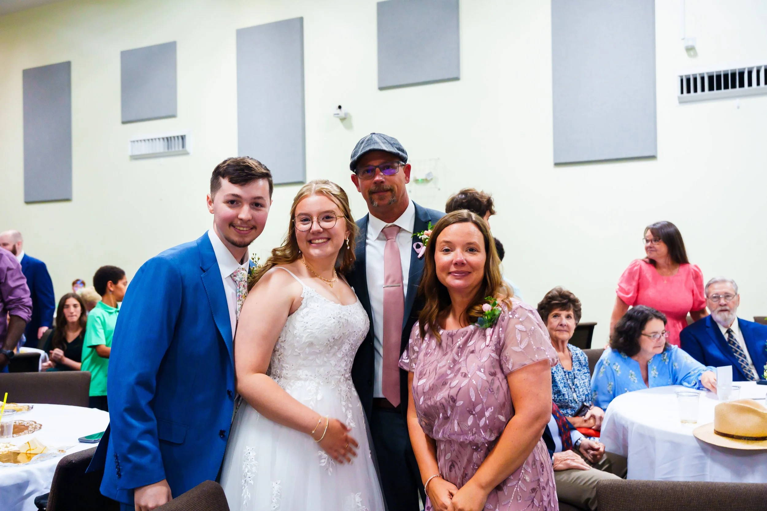 A group of five people celebrating at a wedding reception with friends and family in the background, including three women, a man in a suit, and a groom in a wedding dress.