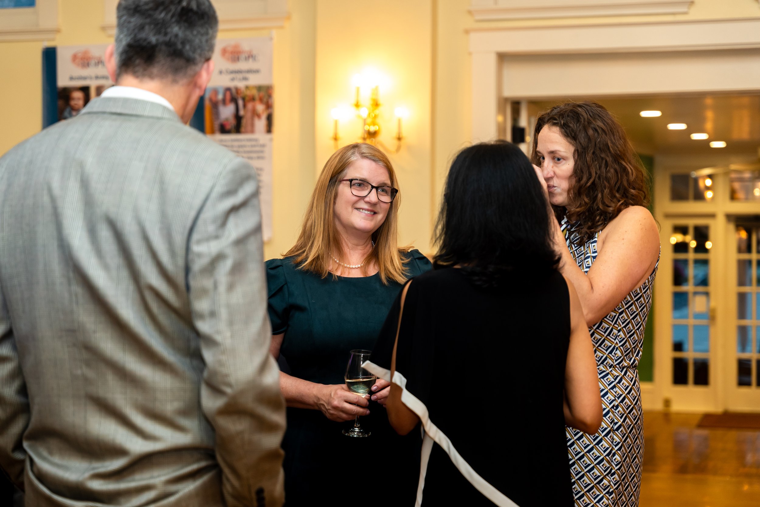 A group of people socializing indoors at a formal event. One woman is holding a glass of wine, and others are engaged in conversation.