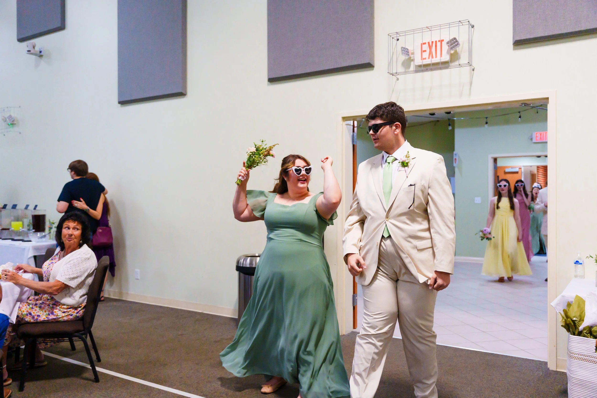 A woman in a green dress and sunglasses is dancing happily at a wedding reception, holding a bouquet of flowers, while a man in a cream-colored suit and sunglasses walks beside her. Other guests are seen in the background, some embracing, and a group