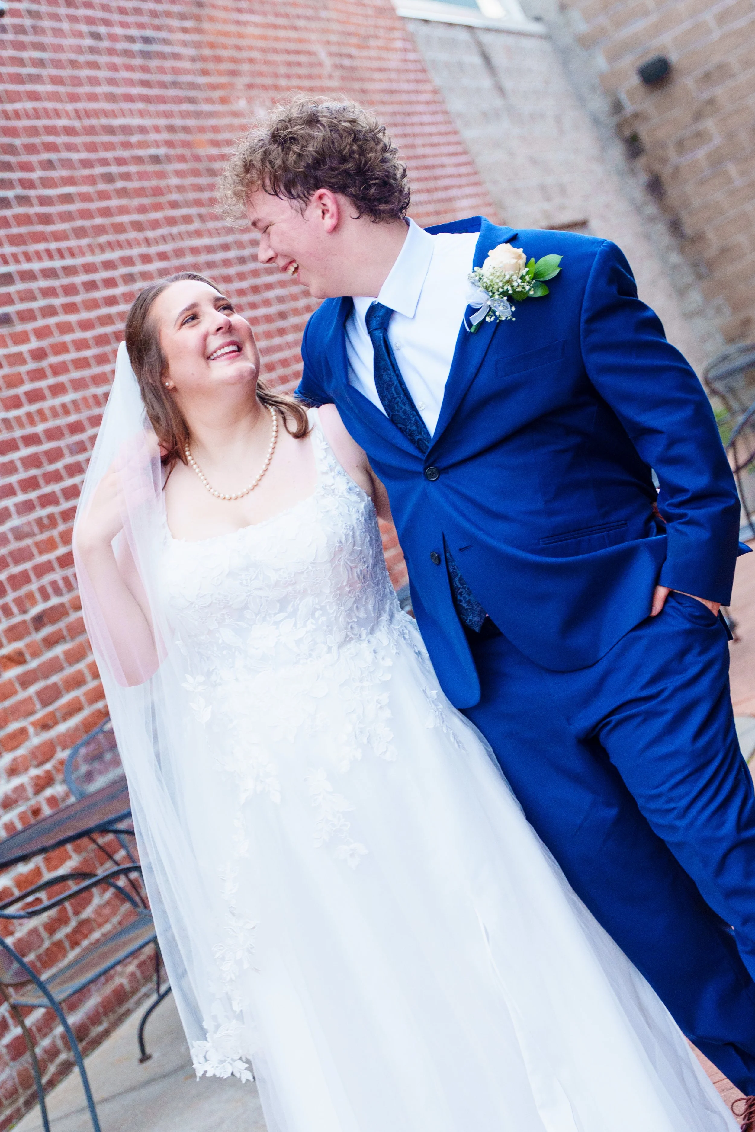 A bride and groom smiling at each other outside, with a brick wall background. The bride is wearing a white wedding dress with lace details and a pearl necklace. The groom is in a blue suit with a white shirt, blue tie, and a boutonniere. They are lo