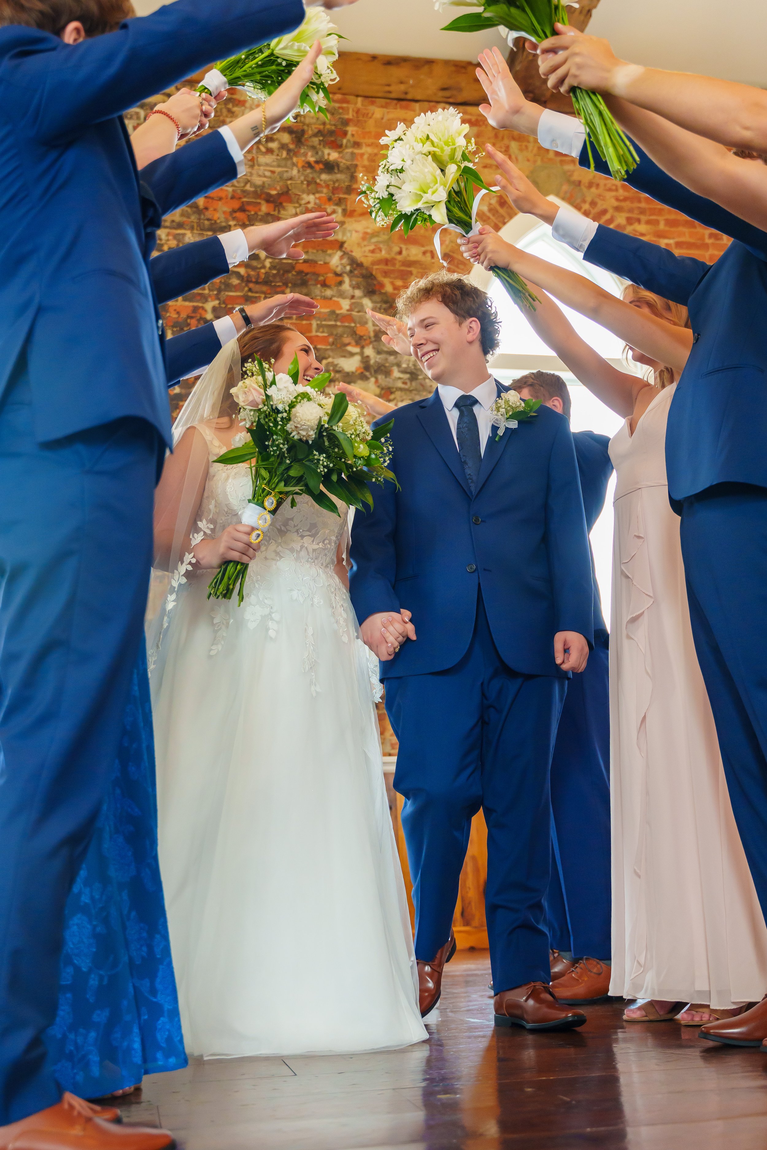 People in formal attire holding bouquets of flowers, forming a corridor. The groom and bride are smiling at each other, surrounded by guests. The setting appears to be indoors with a brick wall and wooden flooring.