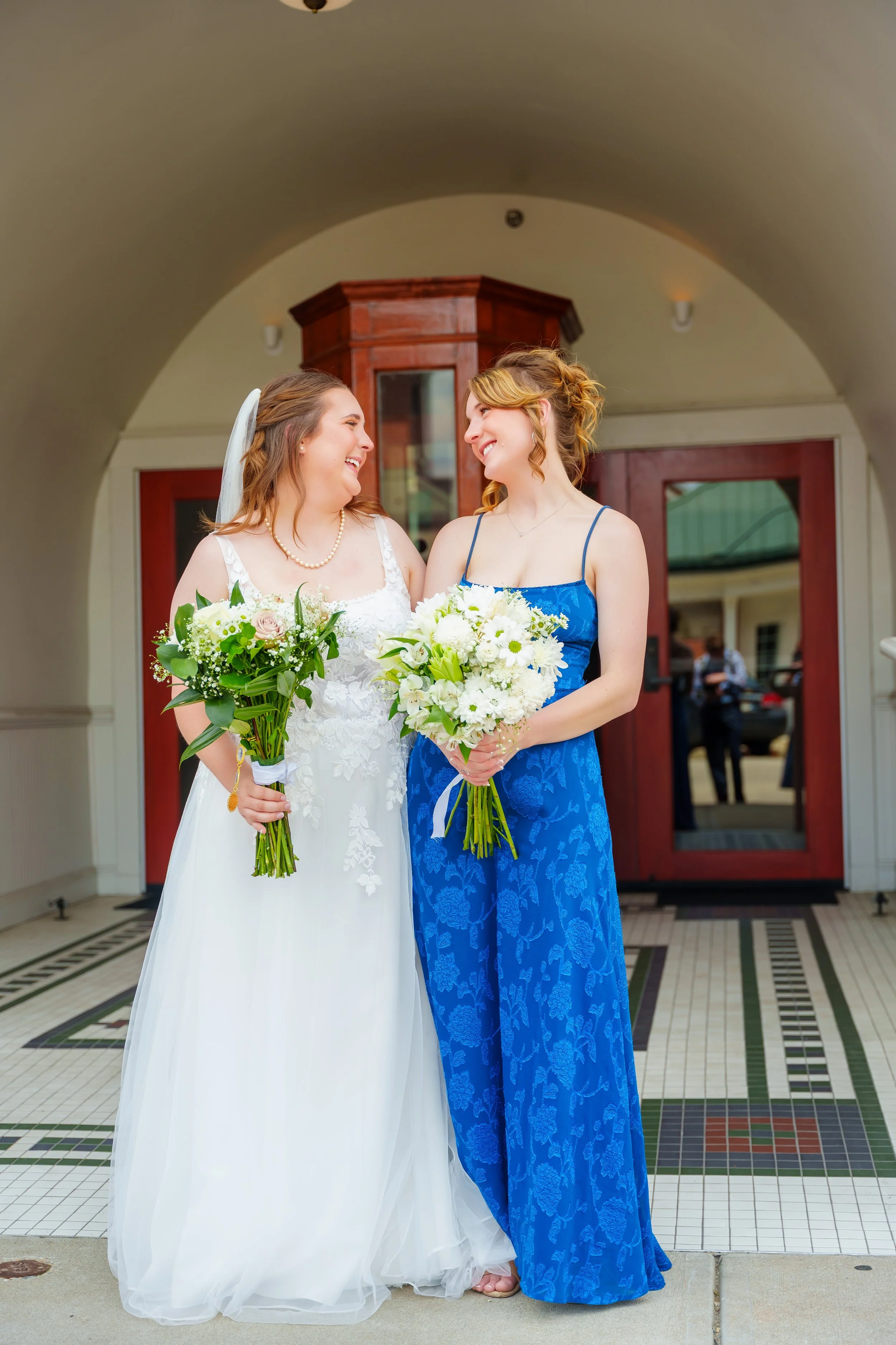 Two women, one in a white wedding dress with a pearl necklace and the other in a blue dress, are smiling and holding bouquets of white and pink flowers, standing in front of a building with a tiled floor and wooden door.