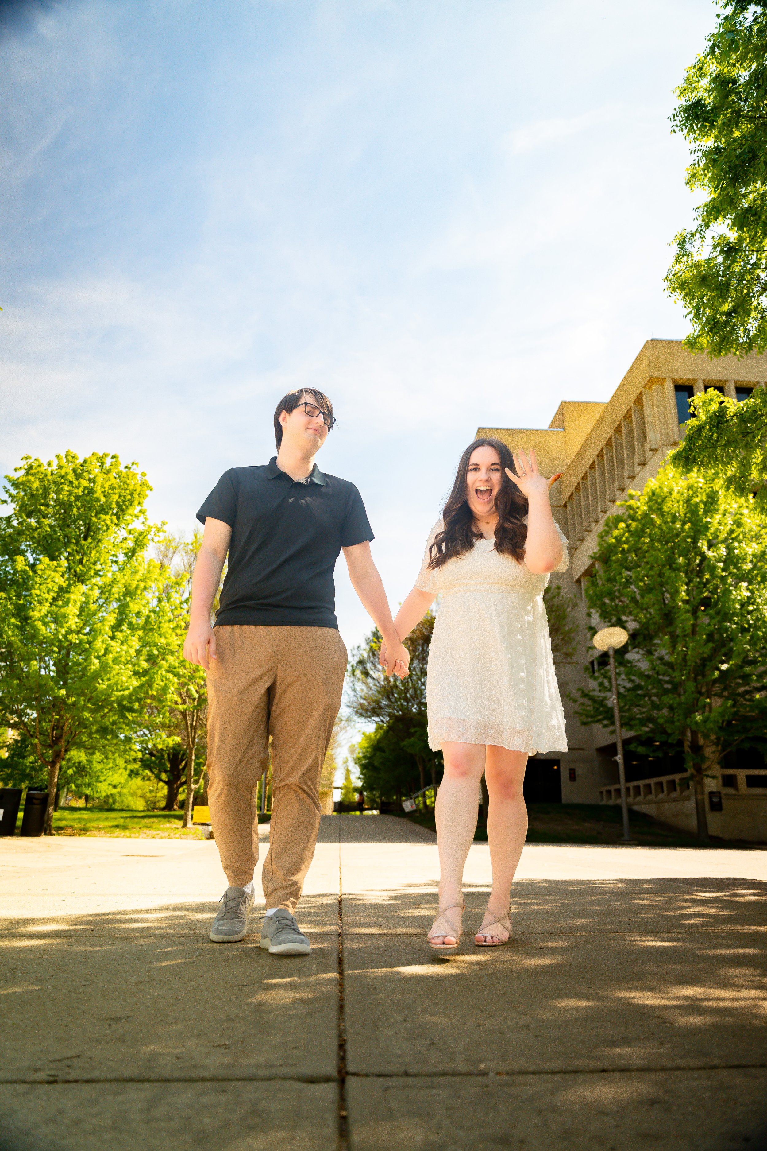 A young couple holding hands and smiling while walking outdoors on a sunny day, surrounded by green trees and a modern building in the background.