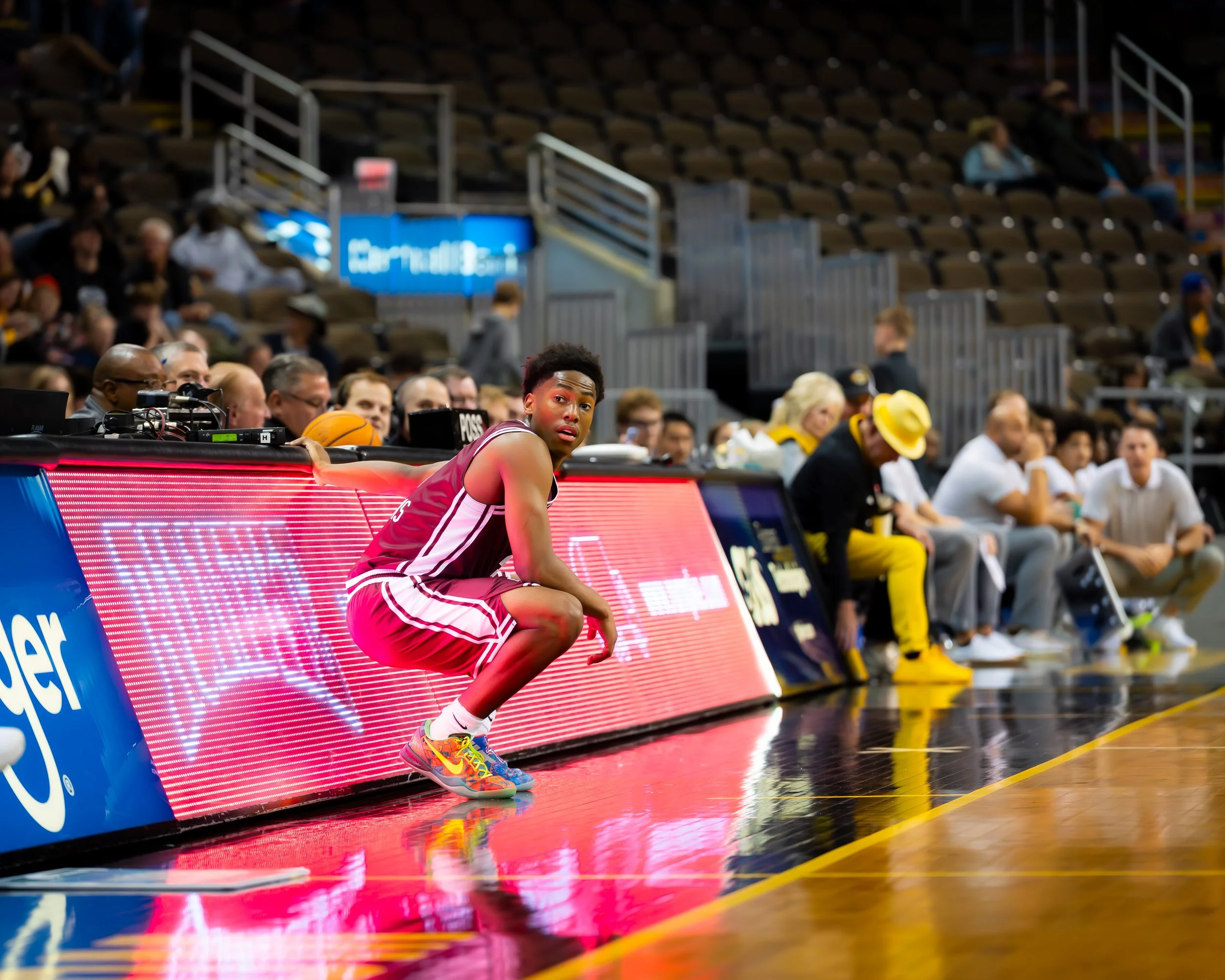 A young male basketball player in a red and white uniform crouches on the sideline of a basketball court, looking towards the camera. The background features a row of seated spectators and team staff sitting on benches, with some engaged in conversat