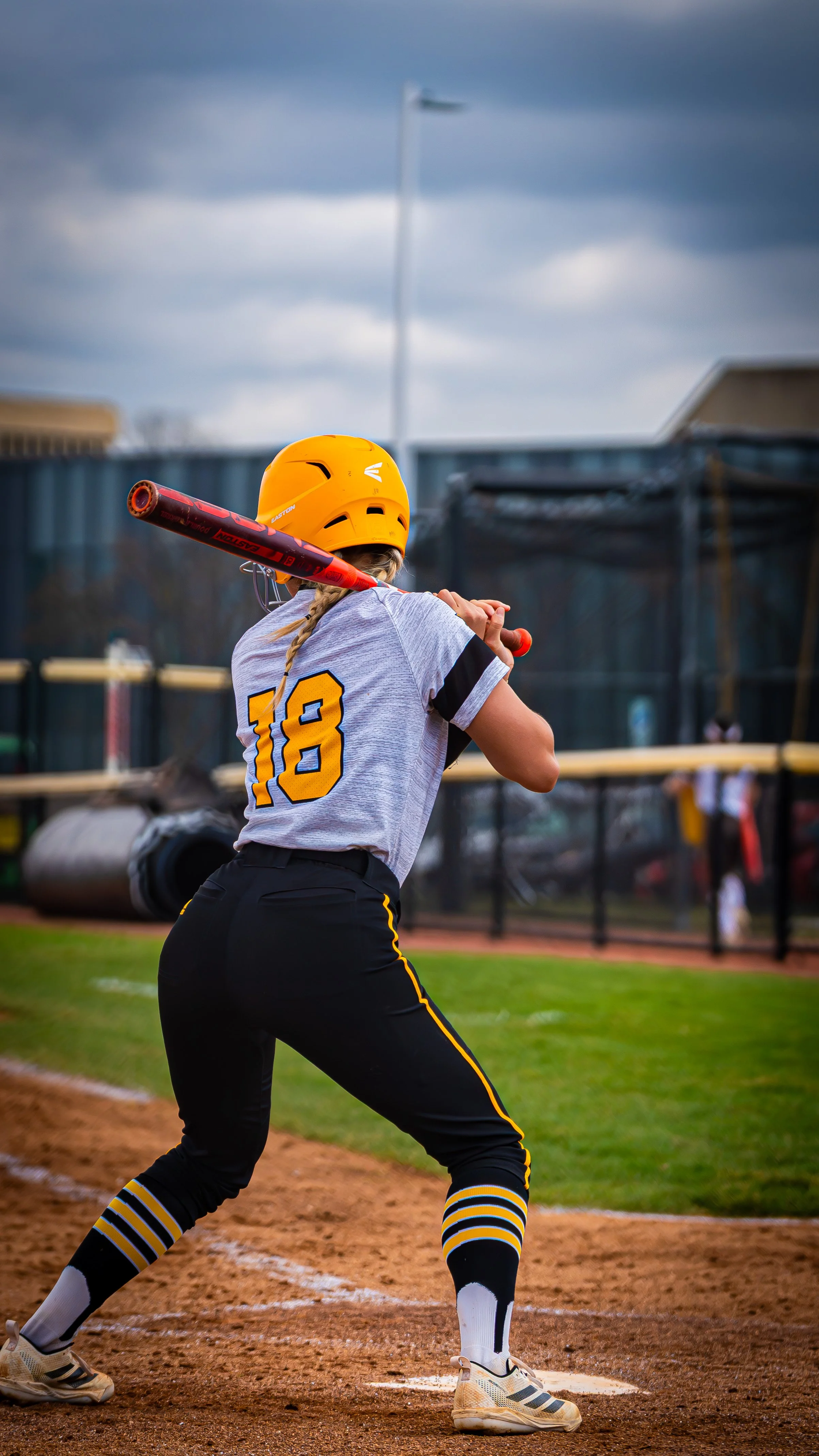A female softball player wearing a yellow helmet and a gray uniform with number 18, standing at bat and ready to hit the ball on a softball field during an overcast day.