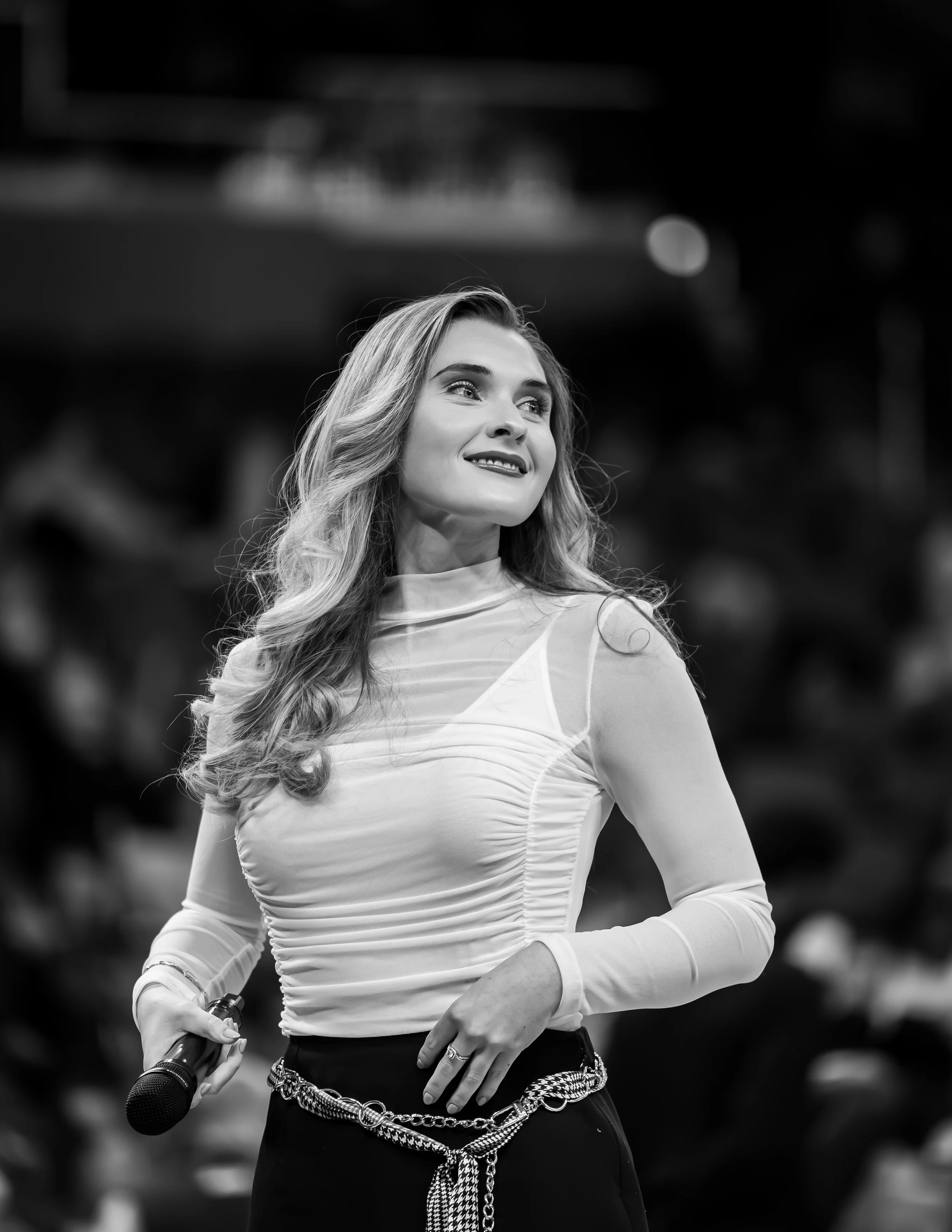 A woman with long, wavy hair wearing a light-colored, long-sleeve top and a chain belt, holding a microphone in her right hand, looking to her left with a smile.