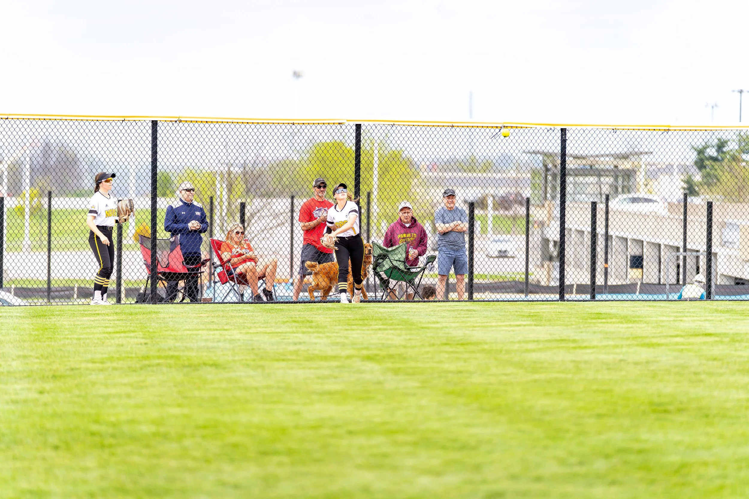 Women in softball uniforms standing near a chain-link fence watching a game, with spectators sitting on chairs behind the fence, a grassy field, and a cloudy sky.