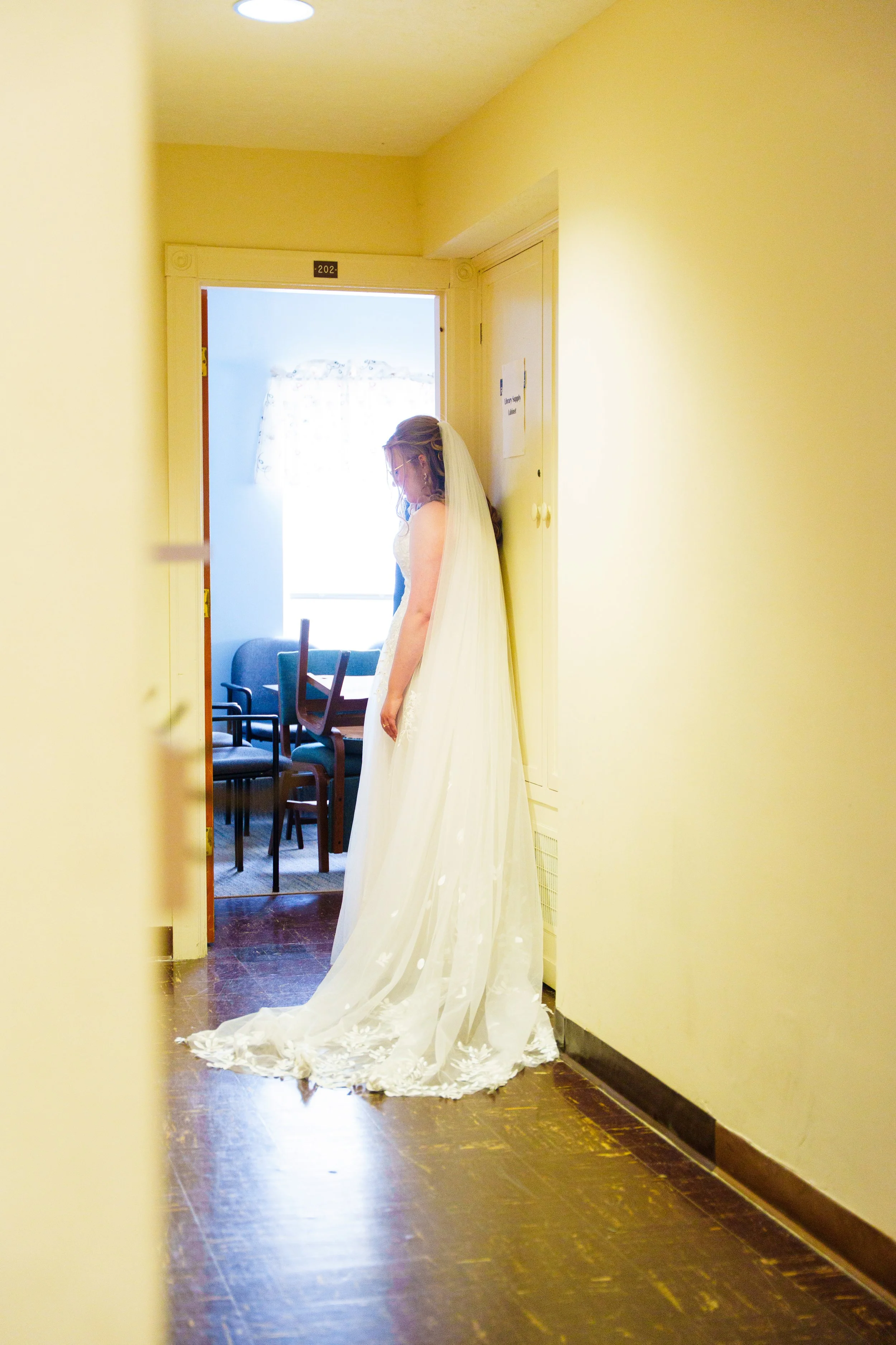 A bride in a wedding dress standing in a doorway, looking down, with her veil flowing behind her.