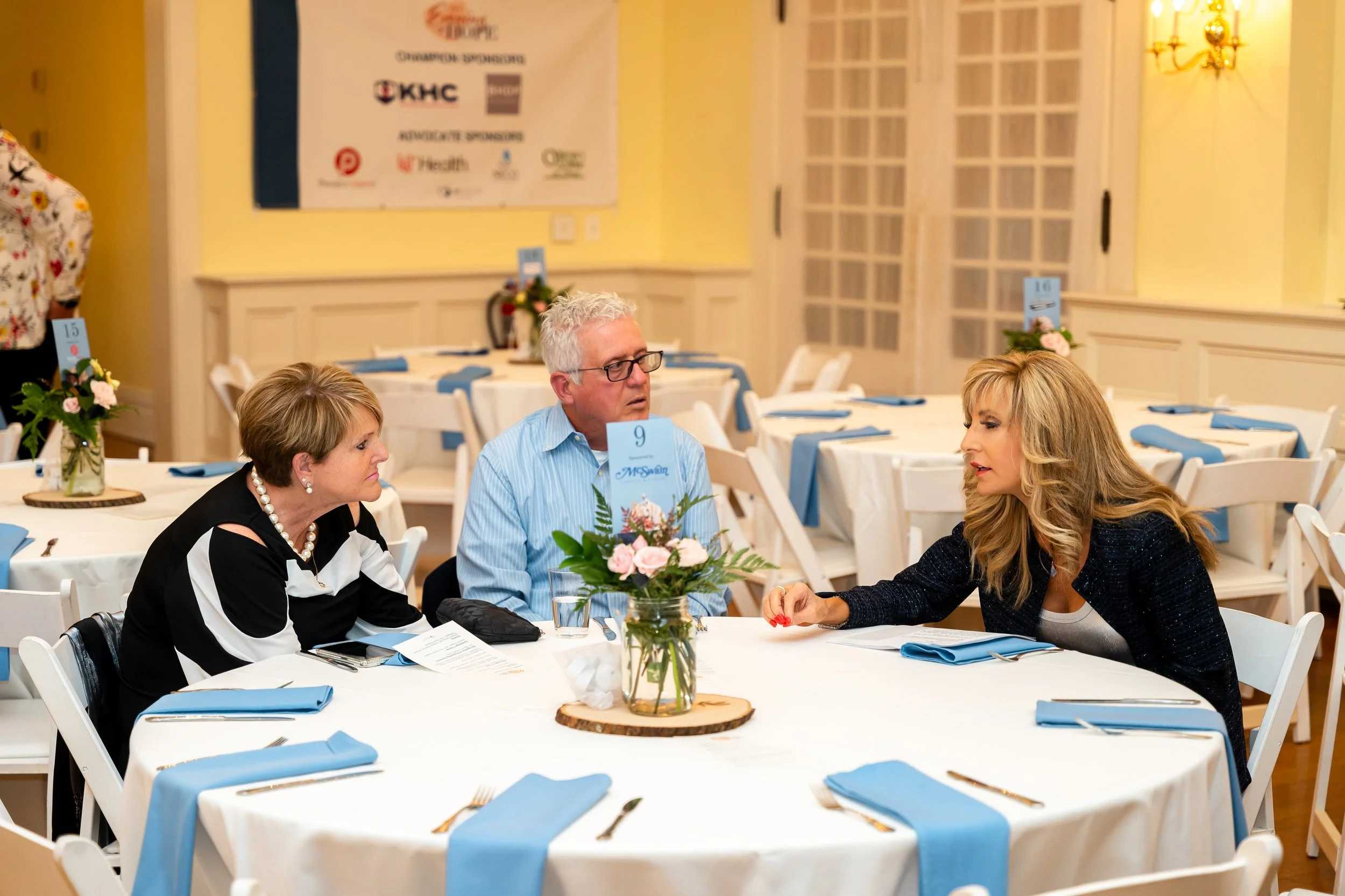 People sitting at decorated banquet tables with blue napkins, floral centerpieces, and event programs, engaged in conversation at a formal indoor event.