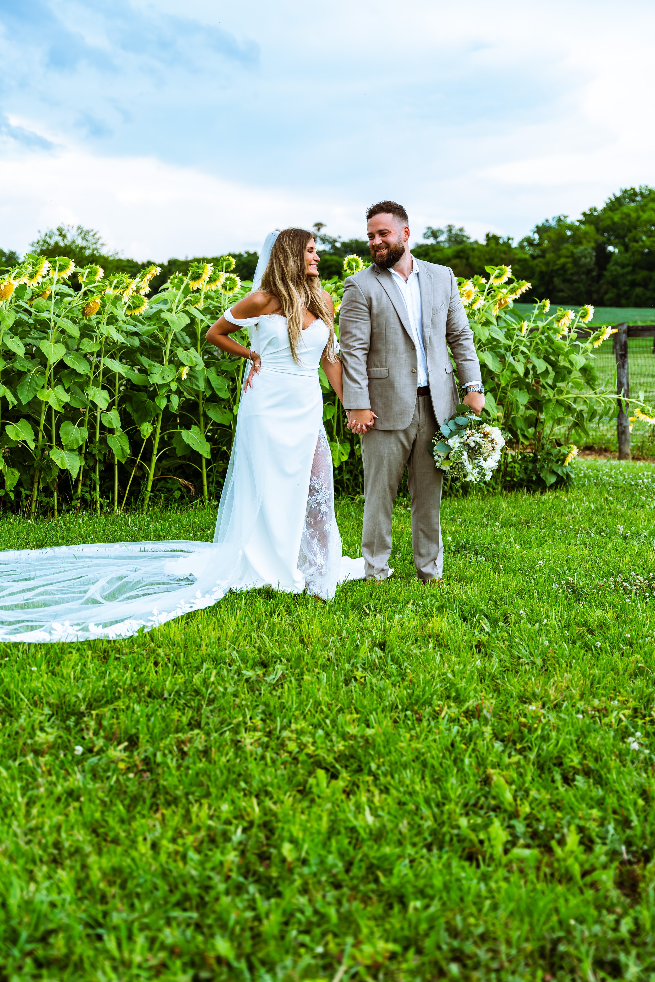 A bride and groom holding hands outdoors in a lush green field with sunflowers, during daytime, with a partly cloudy sky.