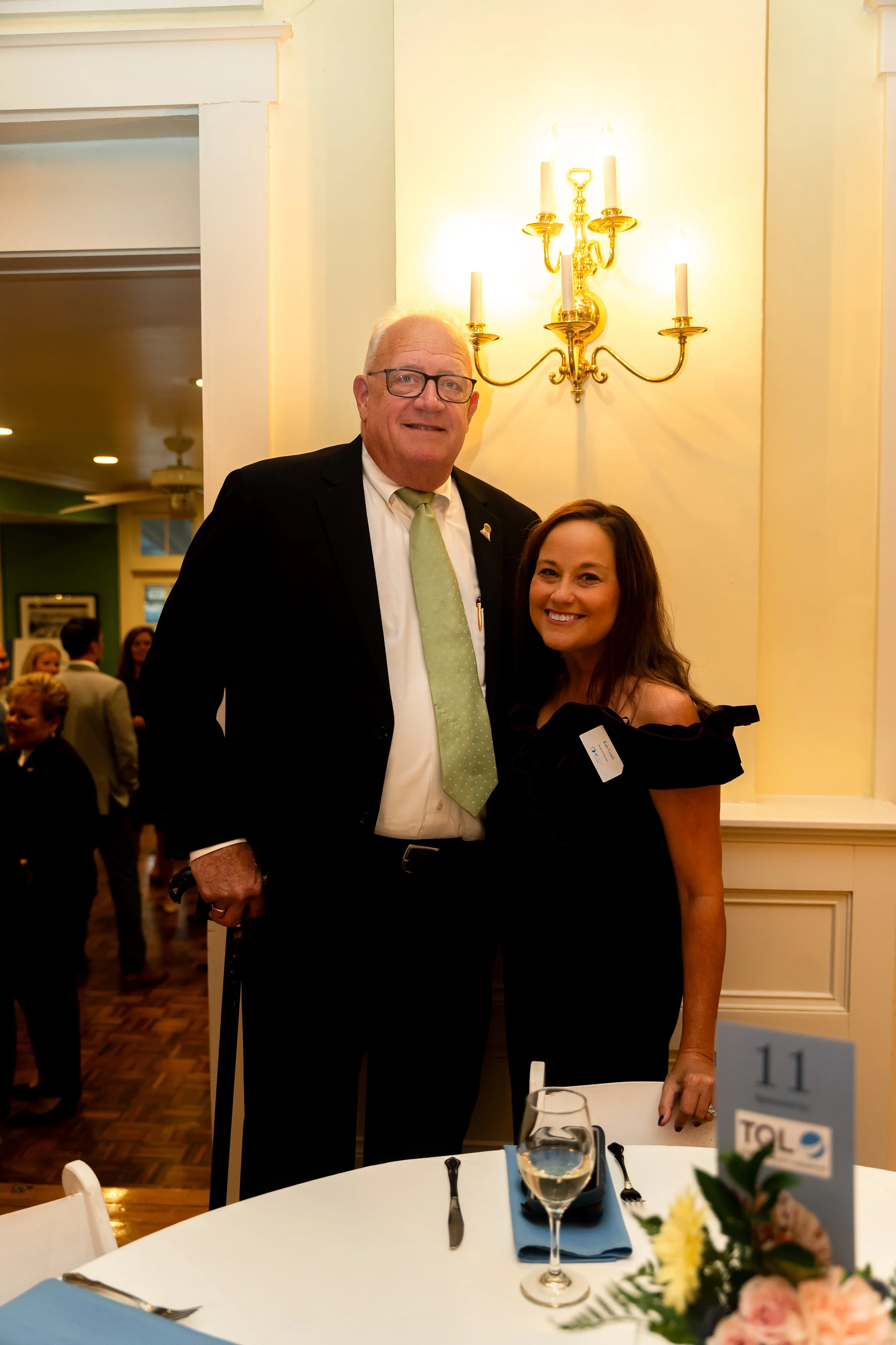 A man and woman smiling and posing together at a formal event, standing behind a table with a blue napkin, silverware, a wine glass, and a floral centerpiece. The man is wearing a black suit, white shirt, and light green tie, and the woman is wearing