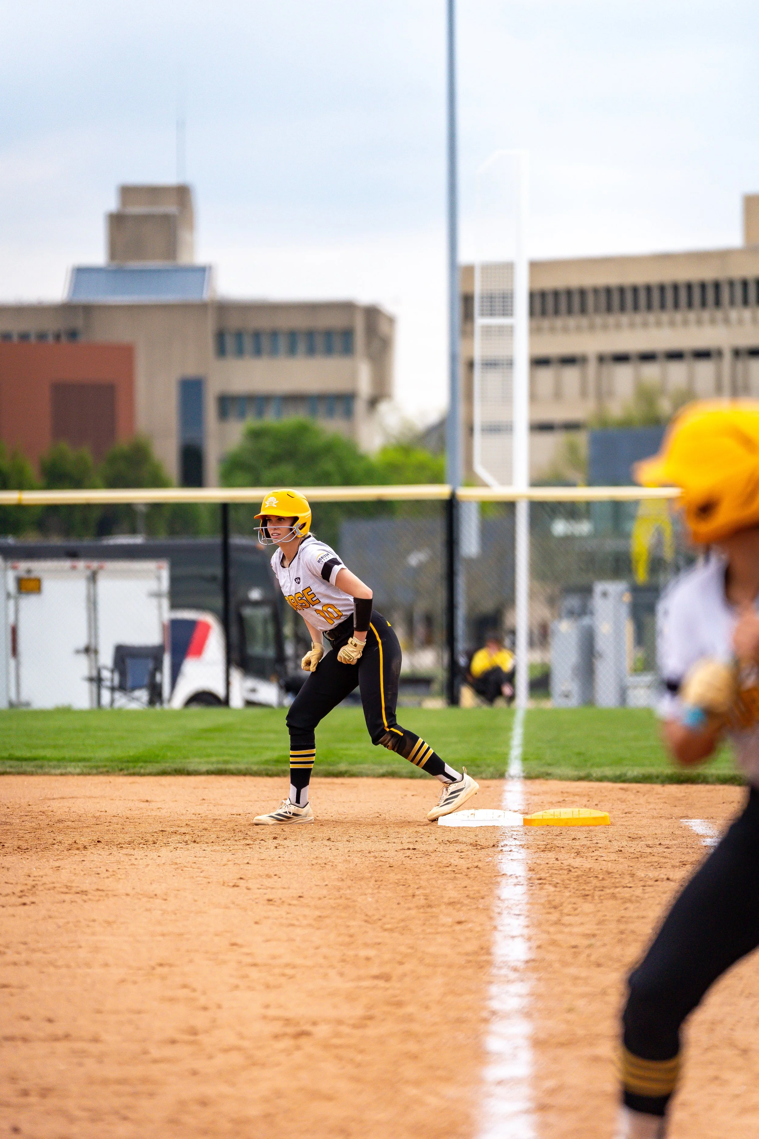 A female softball player stands on base during a game, wearing a yellow helmet, black pants with yellow stripes, and a white jersey with yellow accents. The field has a reddish clay infield and green grass outfield, with a cityscape of tall buildings