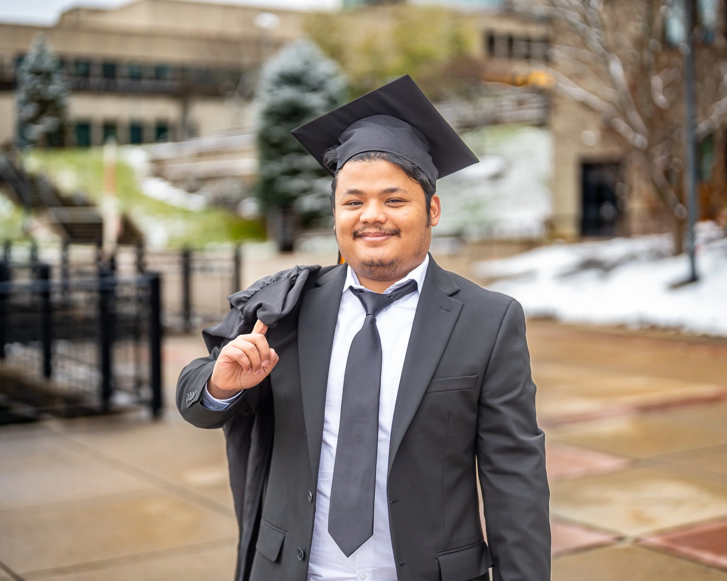 A young man in graduation cap and gown smiling outdoors with buildings and trees in the background.